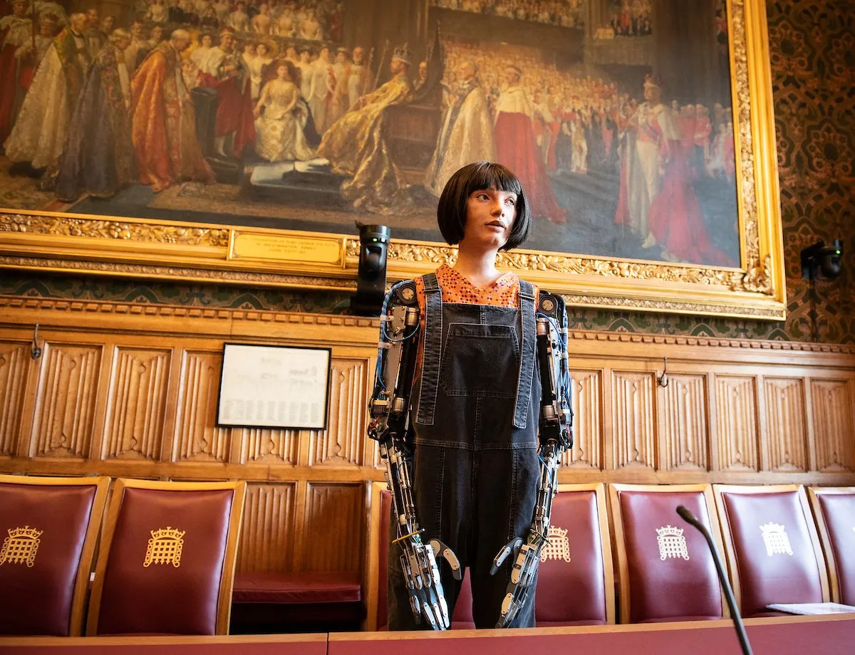 Ai-Da, a humanoid robot with a bobbed haircut, stands in dungarees at a committee table in the House of Lords beneath a large painting.
