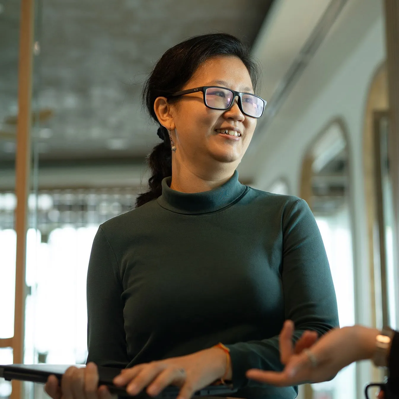Smiling woman with glasses in a dark green turtleneck holds a laptop and gestures while speaking in a bright, blurred indoor setting.
