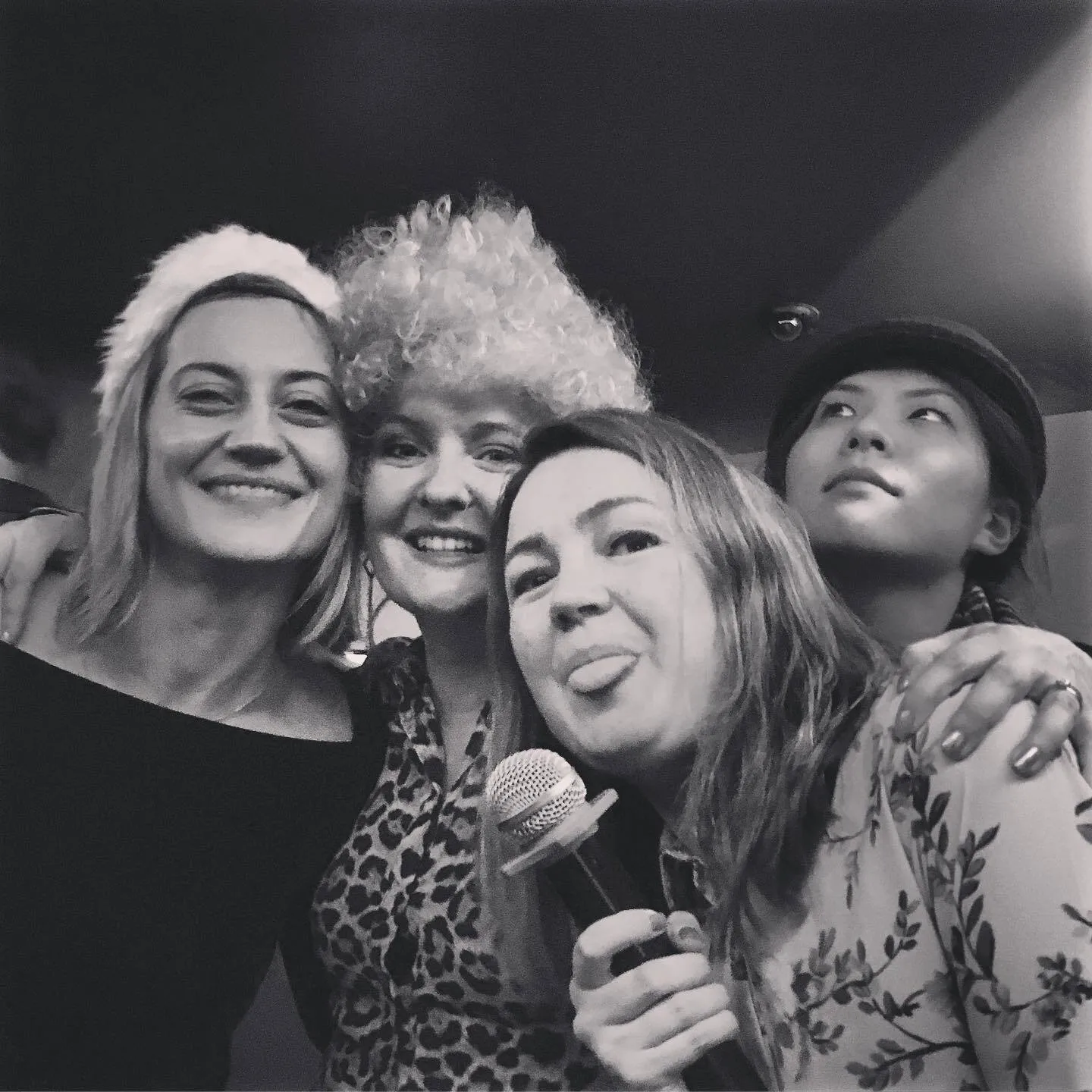 A black-and-white selfie of four women, one in a Santa hat and one in a fluffy wig, another holding a microphone and sticking out her tongue.