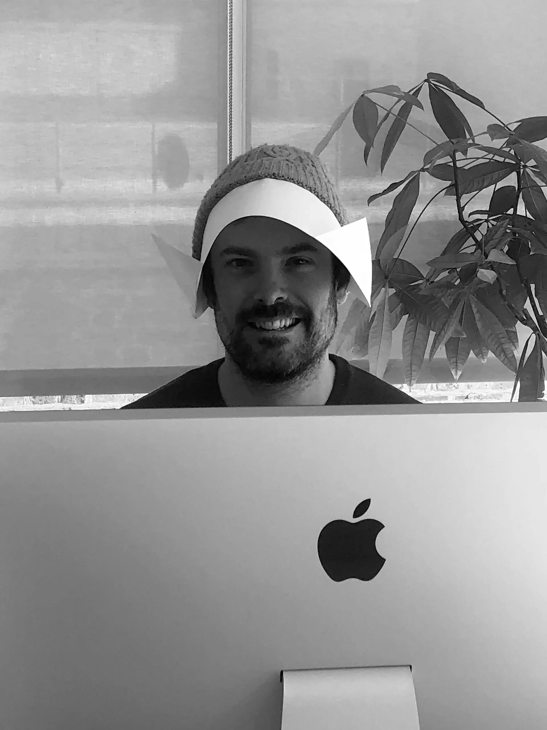 Black and white photo of a smiling man in a beanie and a paper hat, peering over the top of an iMac beside a houseplant.
