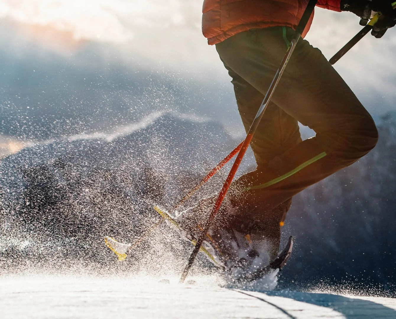 A skier in an orange jacket and brown pants carves through snow, kicking up a bright spray of powder in backlit sunshine, with ski poles and skis visible in mid-turn.