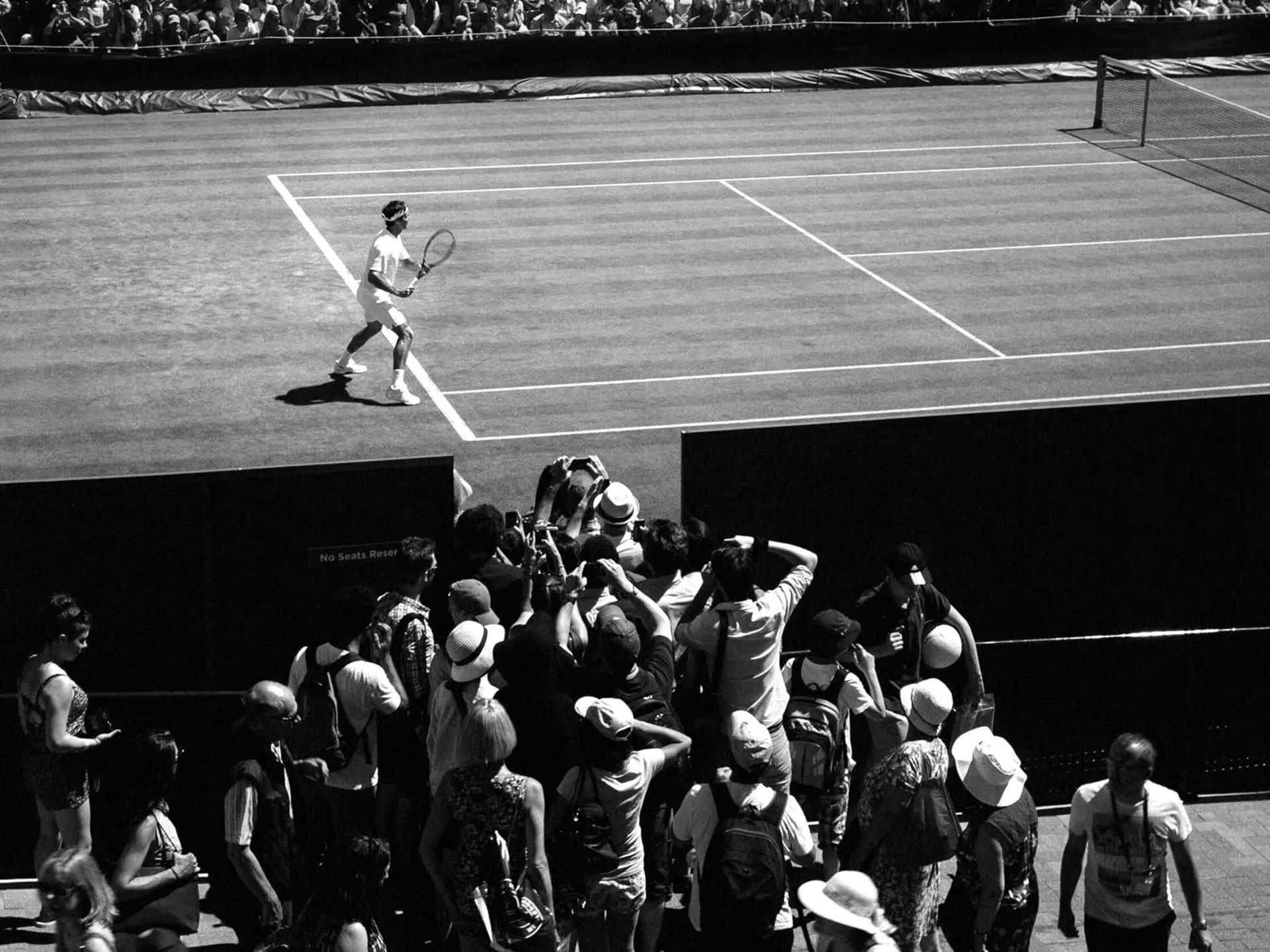 Black and white photograph showing a tennis player in white attire on a grass court, viewed from above, with a crowd of spectators gathered at an opening in the barrier below to watch and take photos.