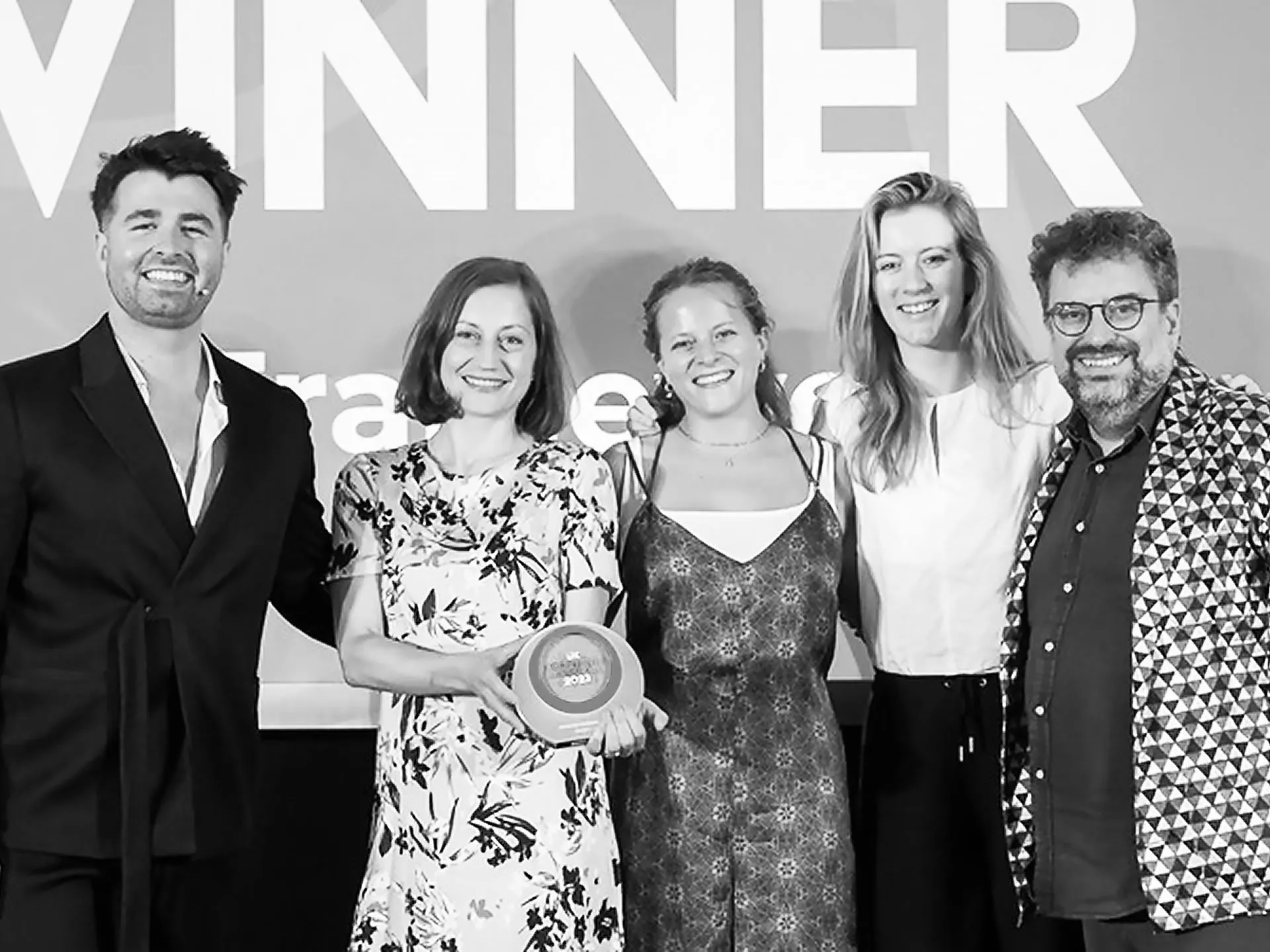 Five smiling people posing together on a stage in front of a background reading WINNER, while one woman holds a circular award.