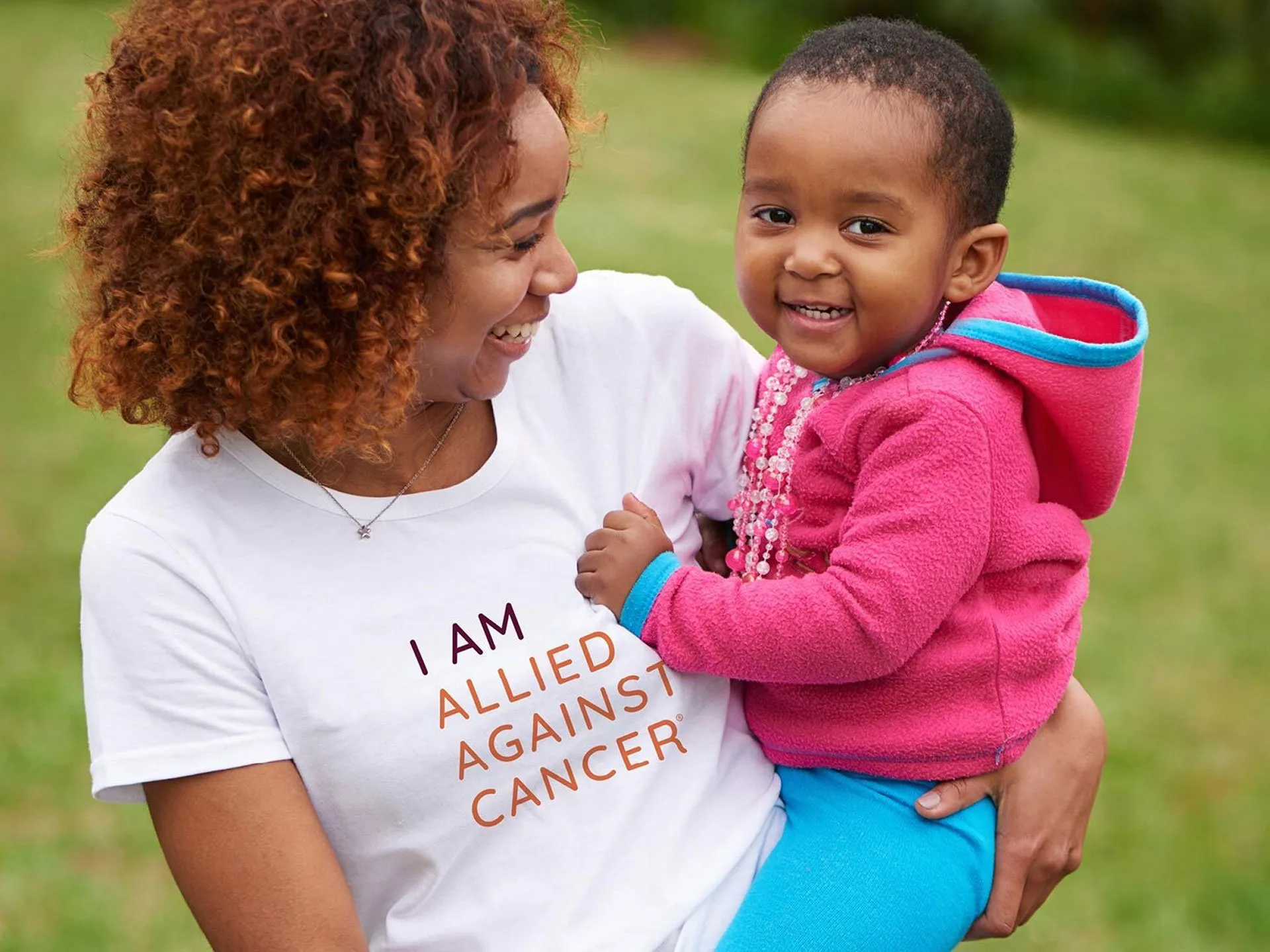 A smiling woman with curly hair holds a cheerful toddler outdoors while wearing a white shirt that reads, "I AM ALLIED AGAINST CANCER."