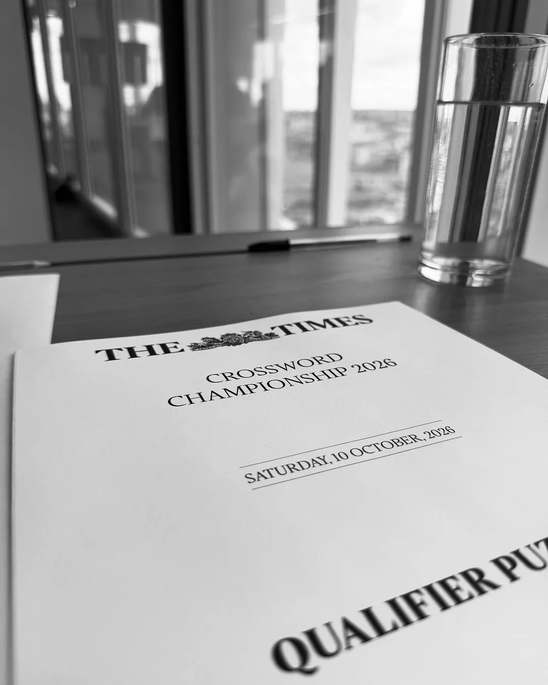 A black and white photo of a printed document titled The Times Crossword Championship 2026 resting on a table, with a glass of water and a pen in the background.