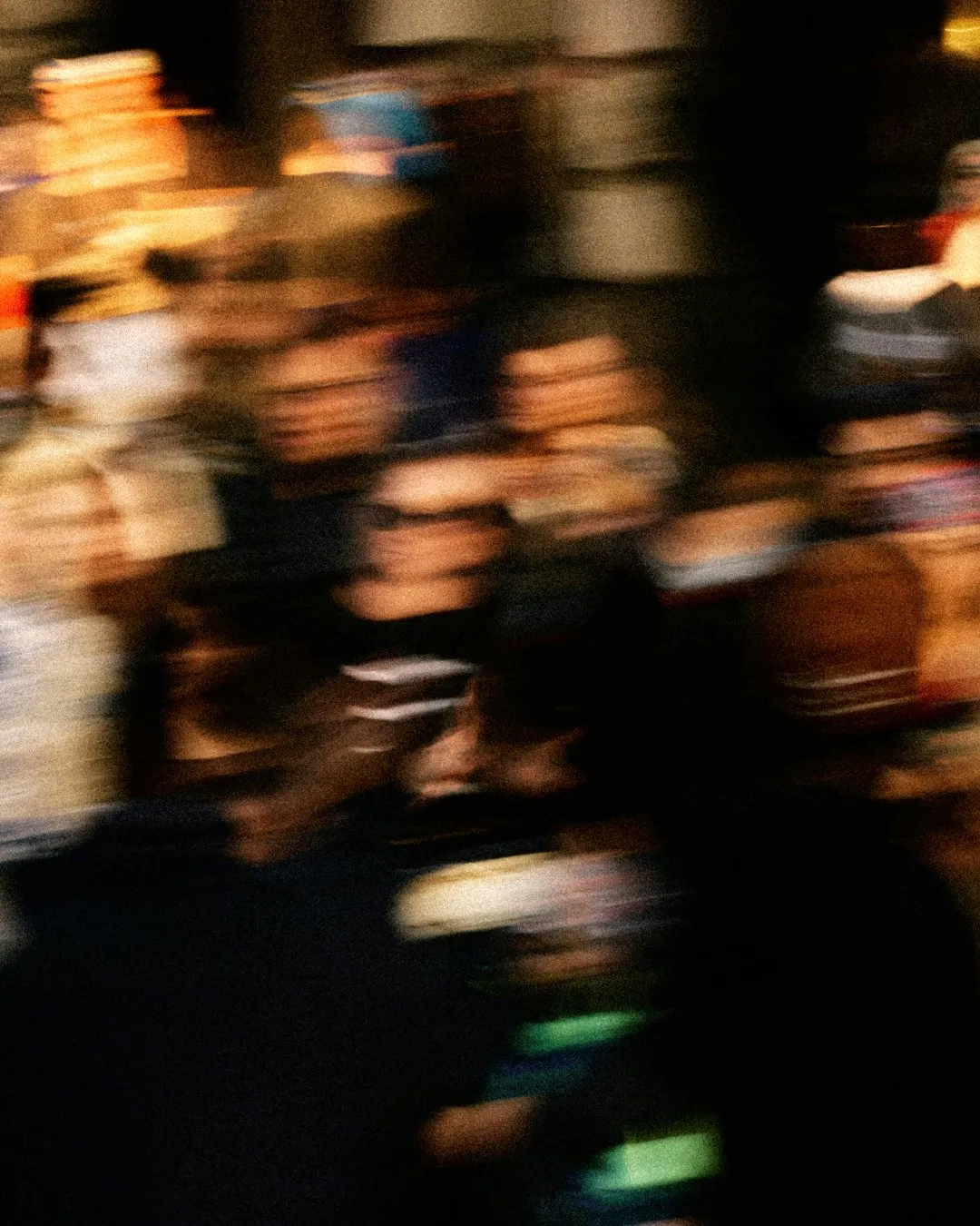 Heavily blurred photo of a crowd of people, with warm amber light streaks and smeared faces against deep shadows.