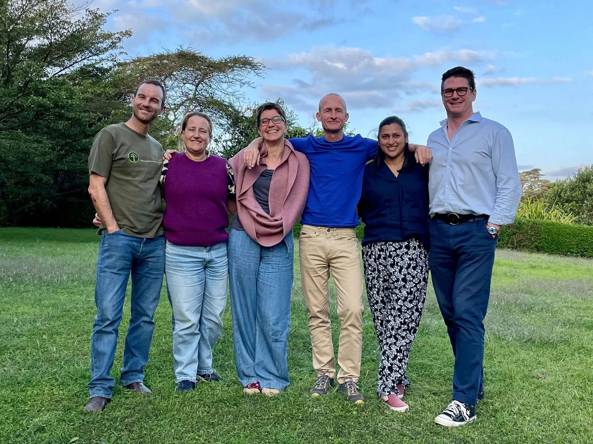 Six smiling adults stand arm-in-arm in a grassy field backed by lush trees and a blue sky.
