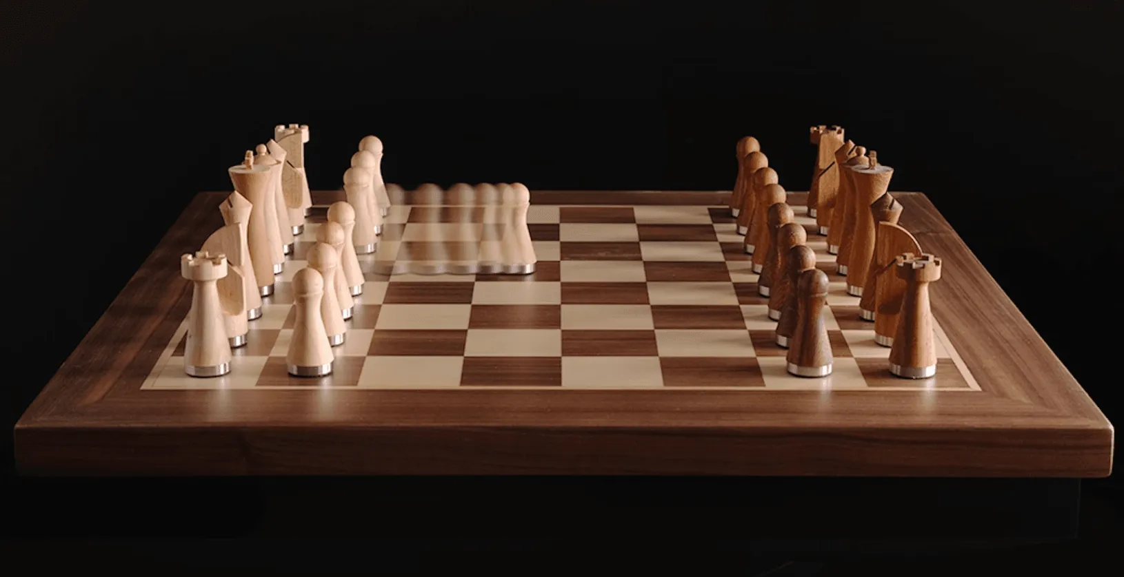 A wooden chessboard with pale and dark carved pieces in their starting positions against a black background, the pawns on one side blurred as if moving.