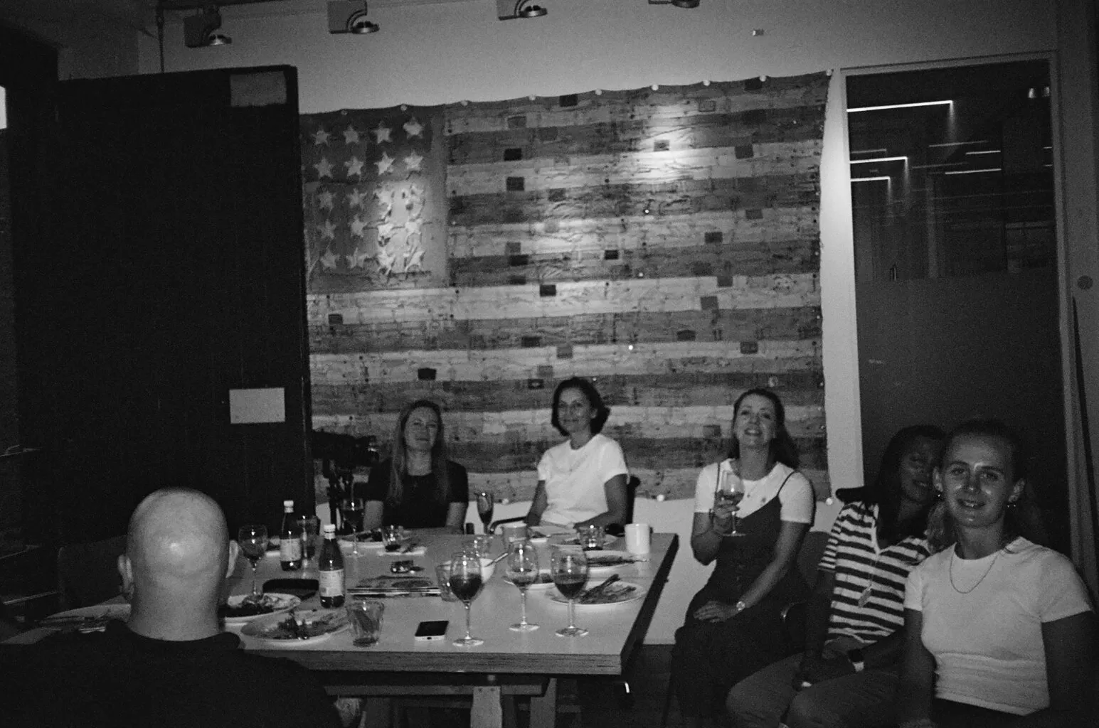 A black-and-white photograph of five colleagues around a studio table with wine glasses, in front of a wall of wooden planks painted as a stars-and-stripes flag.