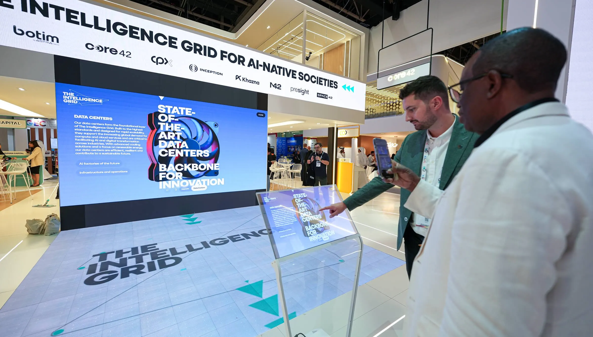 Two men at The Intelligence Grid stand, one pointing at a touchscreen kiosk, in front of a large screen reading 'State-of-the-art data centers: backbone for innovation'.
