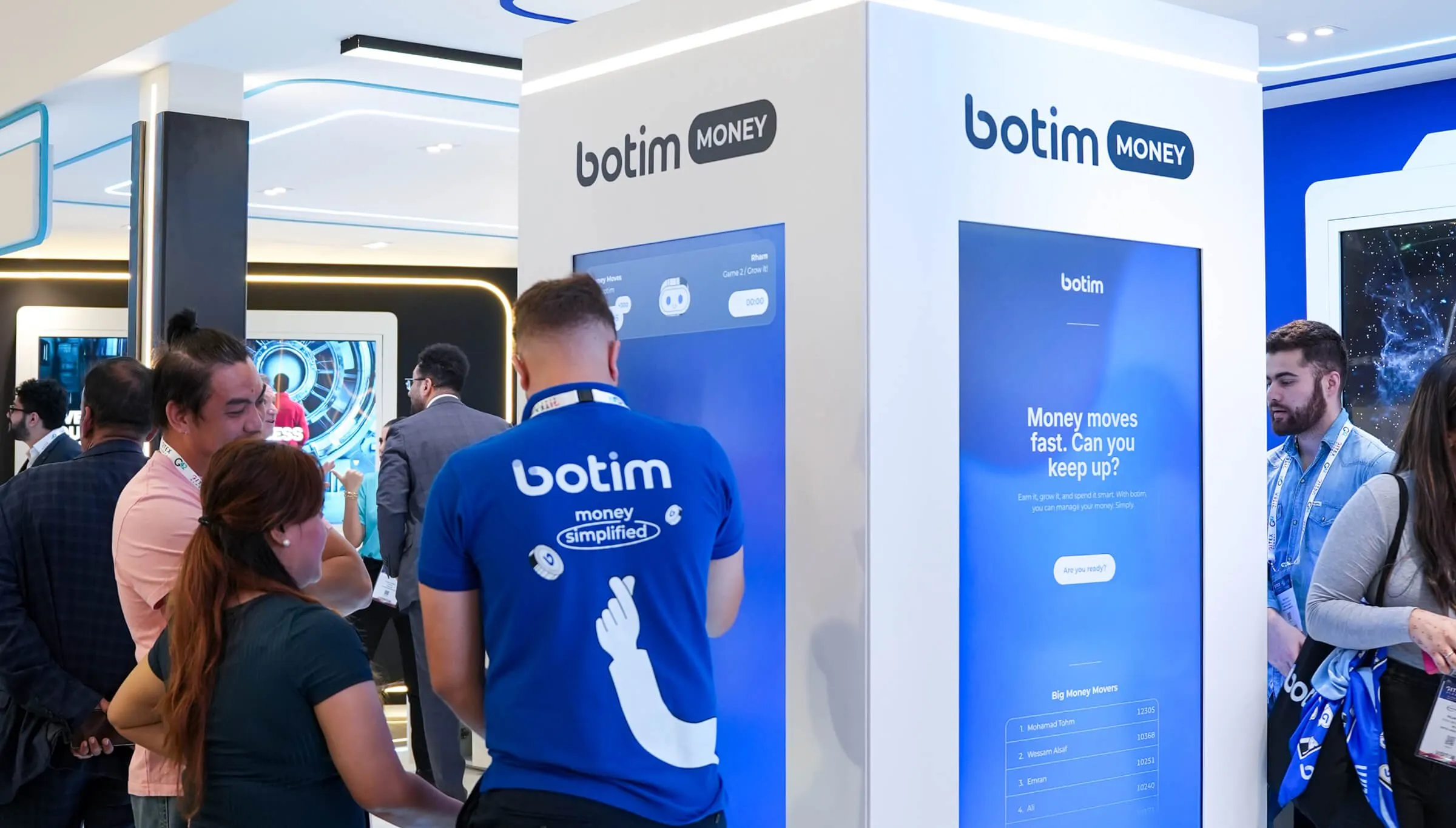 Visitors at the botim Money stand, one in a blue botim T-shirt, in front of a tall screen reading 'Money moves fast. Can you keep up?'