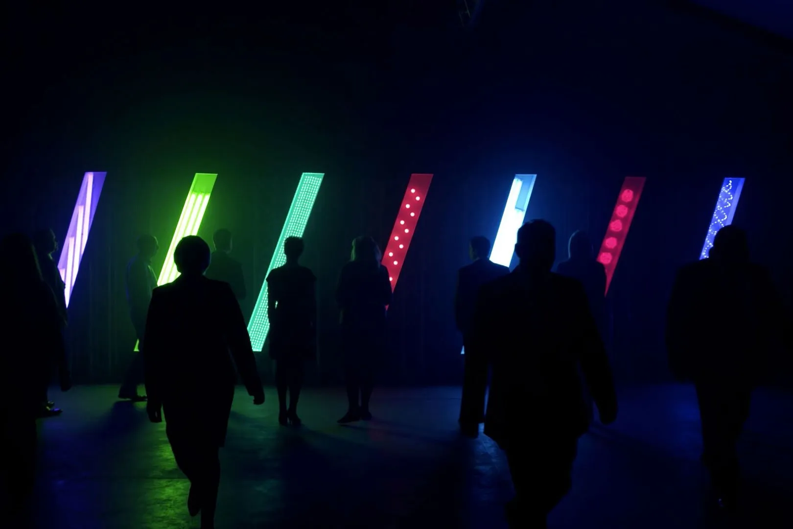 Silhouetted people walking through a dark room past tall, slanted, colorful glowing panels with patterned lights.
