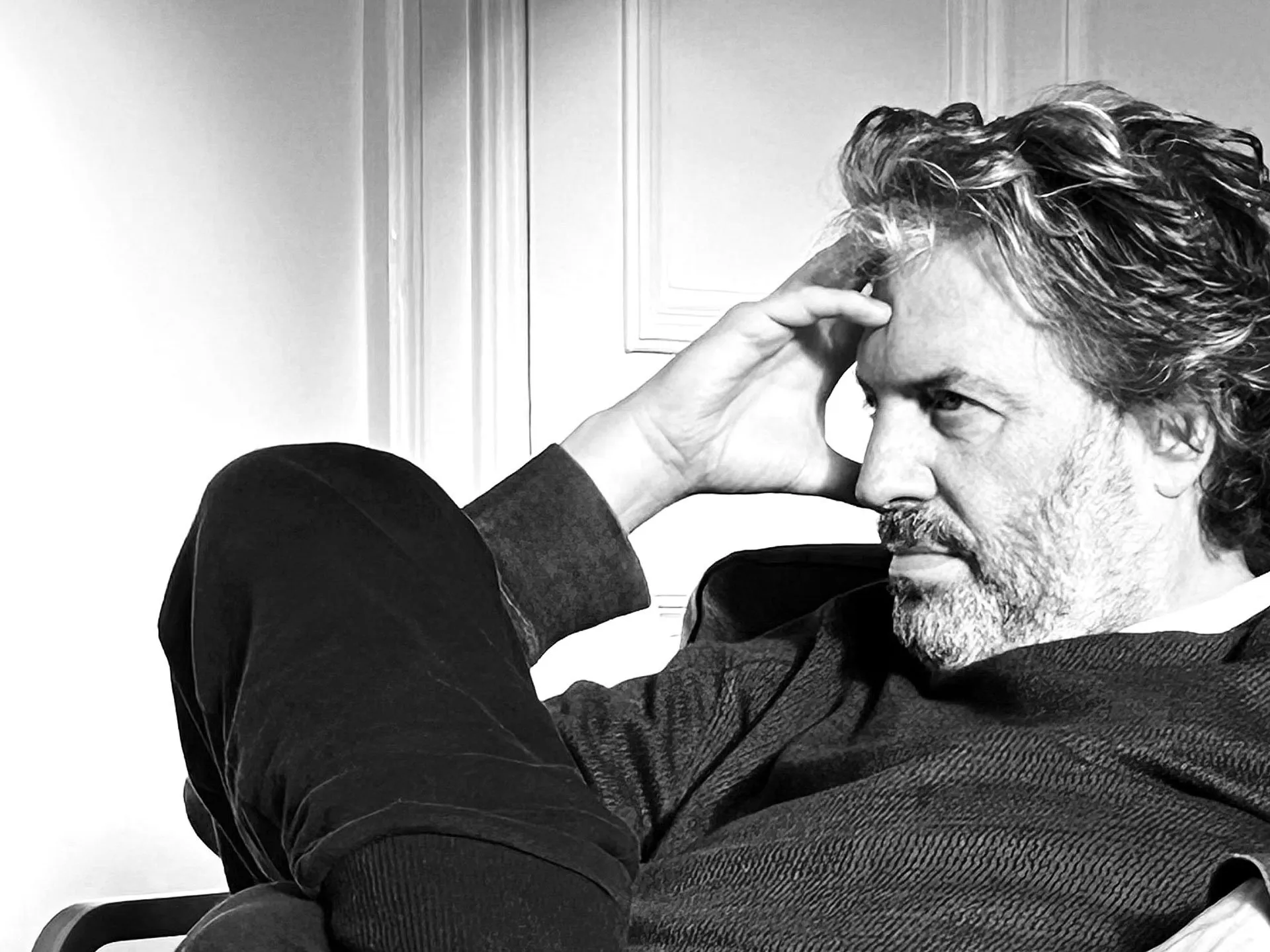 Black and white portrait of a man with wavy graying hair and a beard resting his head on his hand in a thoughtful pose.