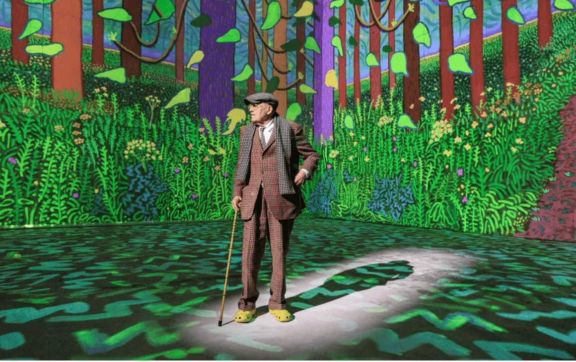 An elderly man in a checked suit and yellow clogs, leaning on a stick, stands in a spotlight in a room filled with projected paintings of a bright green forest.