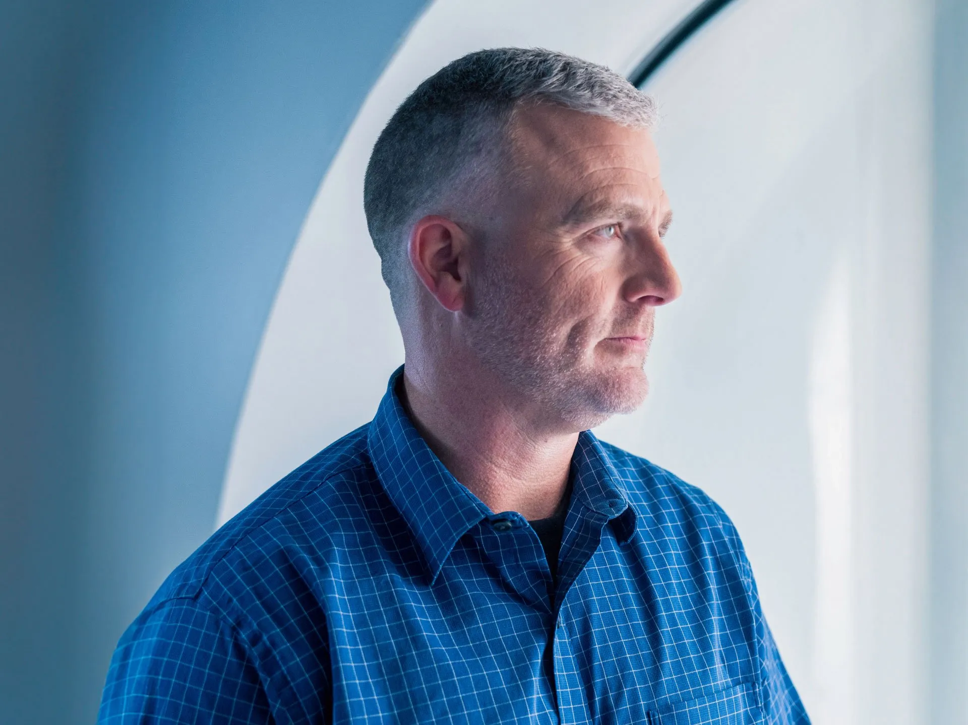 A man with short gray hair wearing a blue checkered shirt looks to the right against a cool-toned background.