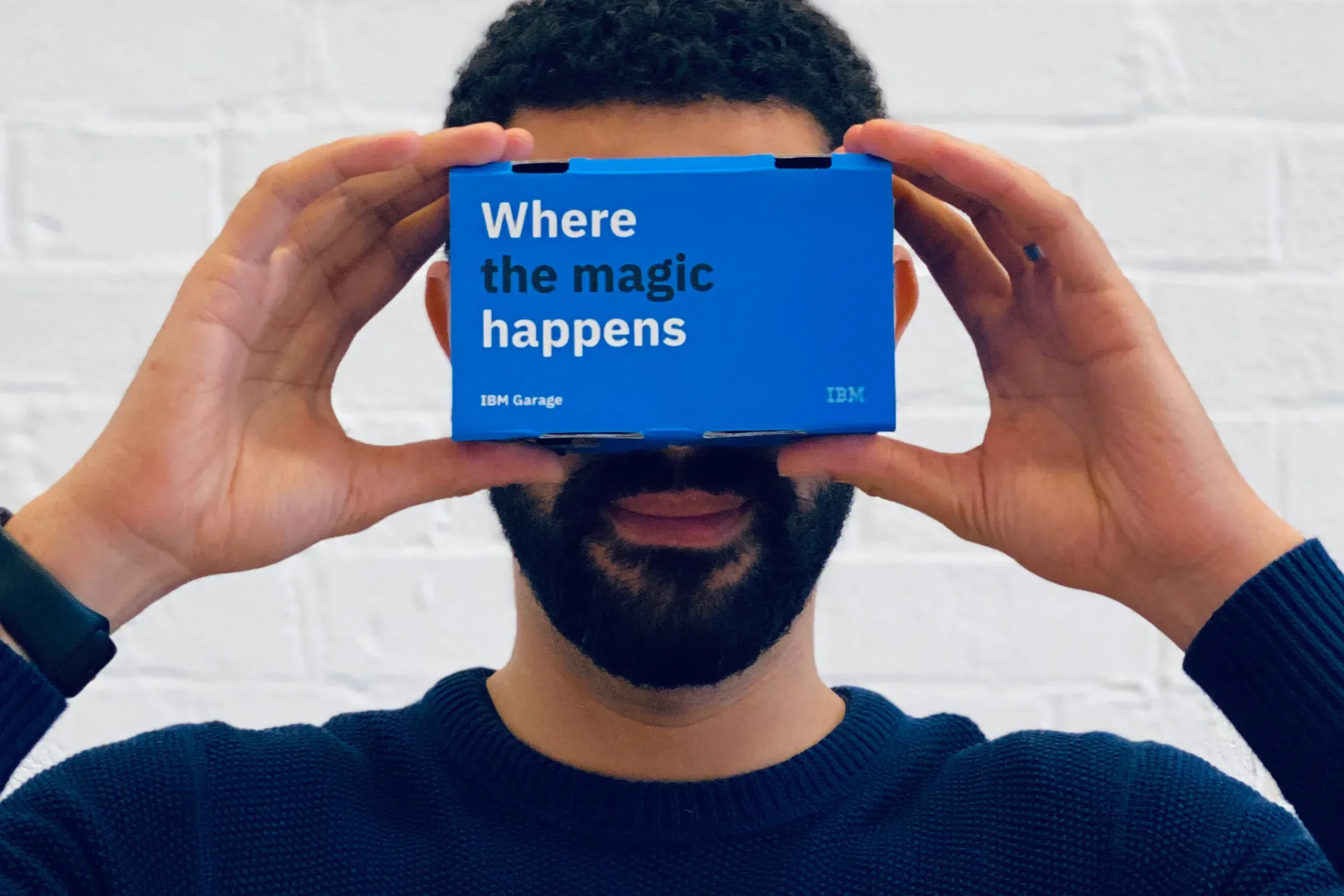 A man holding a blue IBM Garage cardboard VR viewer to his eyes, printed with Where the magic happens