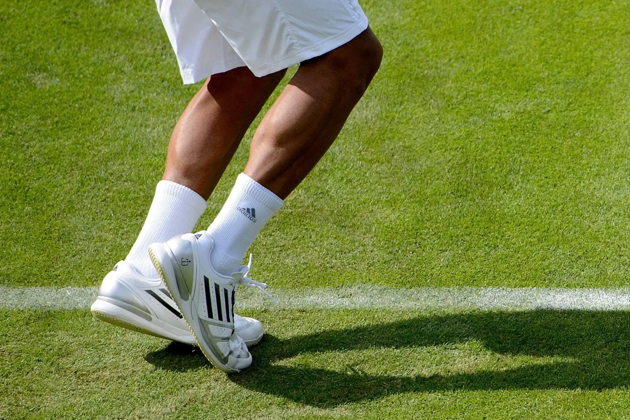 A tennis player's legs and white shoes on a Wimbledon grass court, beside the baseline