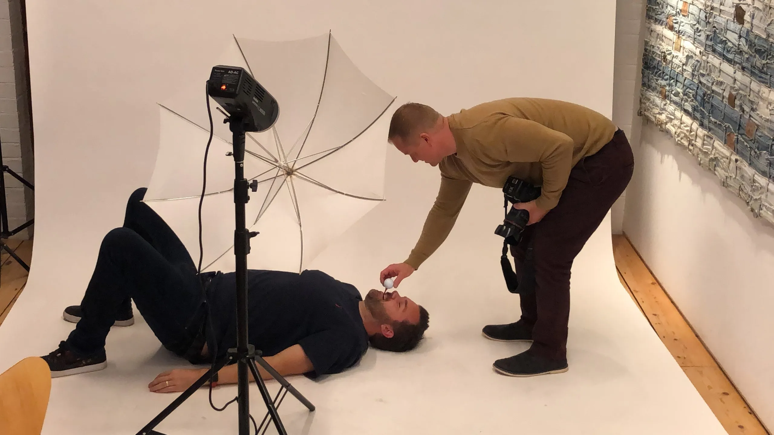 A man lies on his back on the white studio floor while a colleague, camera in hand, leans over him and places something in his mouth; a studio light and umbrella stand beside them.