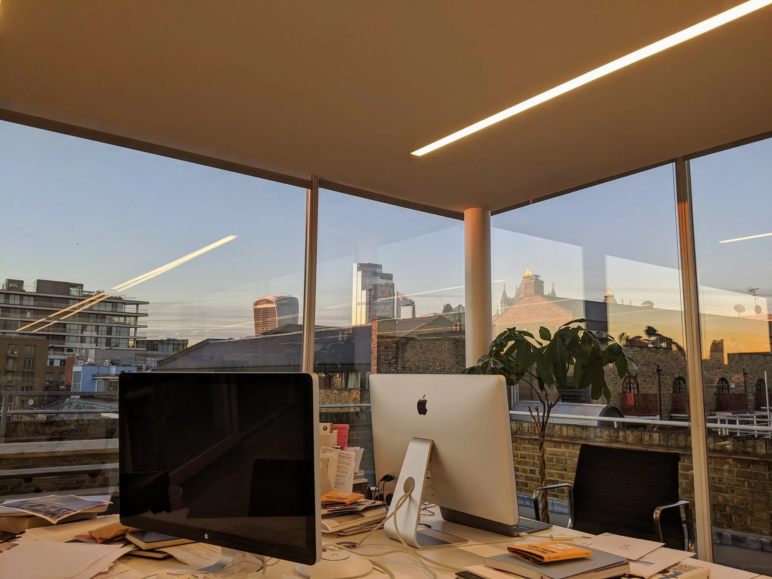 Two Apple screens on a cluttered desk beside floor-to-ceiling windows overlooking the London skyline at dusk.