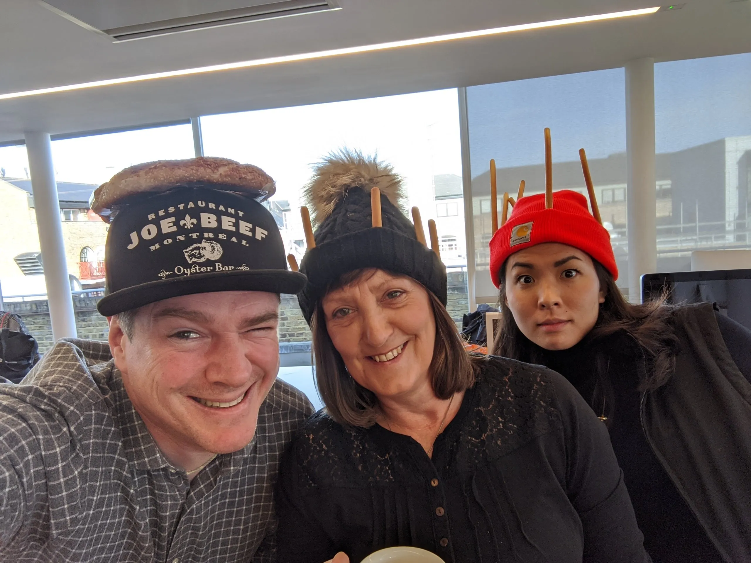 Three colleagues pose for a selfie in food hats: a bread roll on a cap, and two beanies topped with breadsticks.