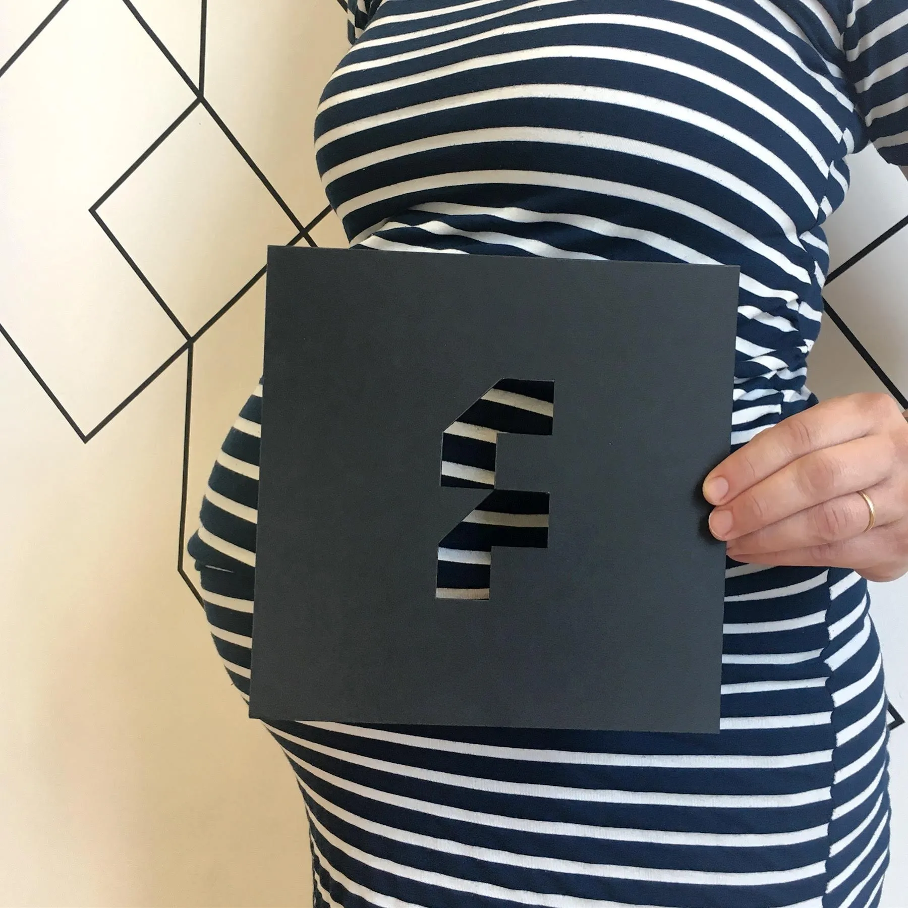 A pregnant woman in a navy-and-white striped dress holds a black cut-out of The Frameworks' F logo in front of her bump.