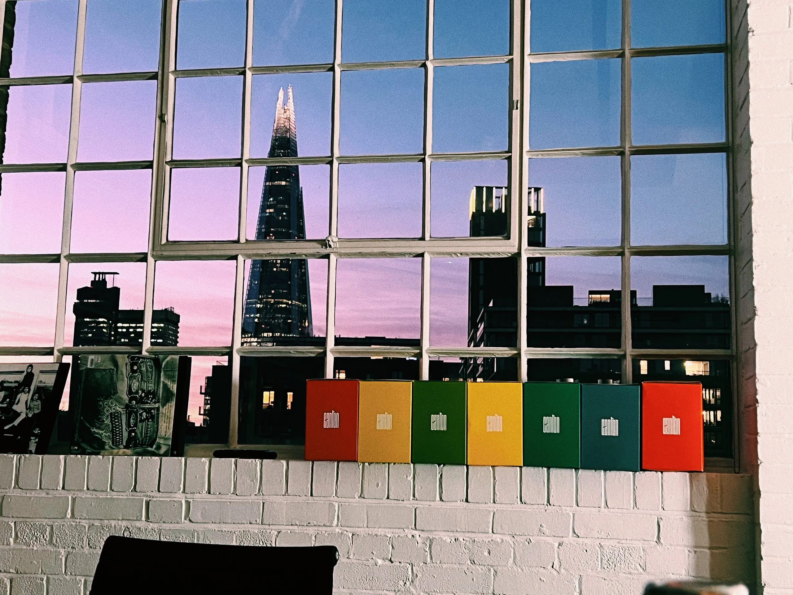 The Shard lit against a pink and lilac dusk sky, seen through a large steel-framed window above a row of coloured boxes on a white brick sill.