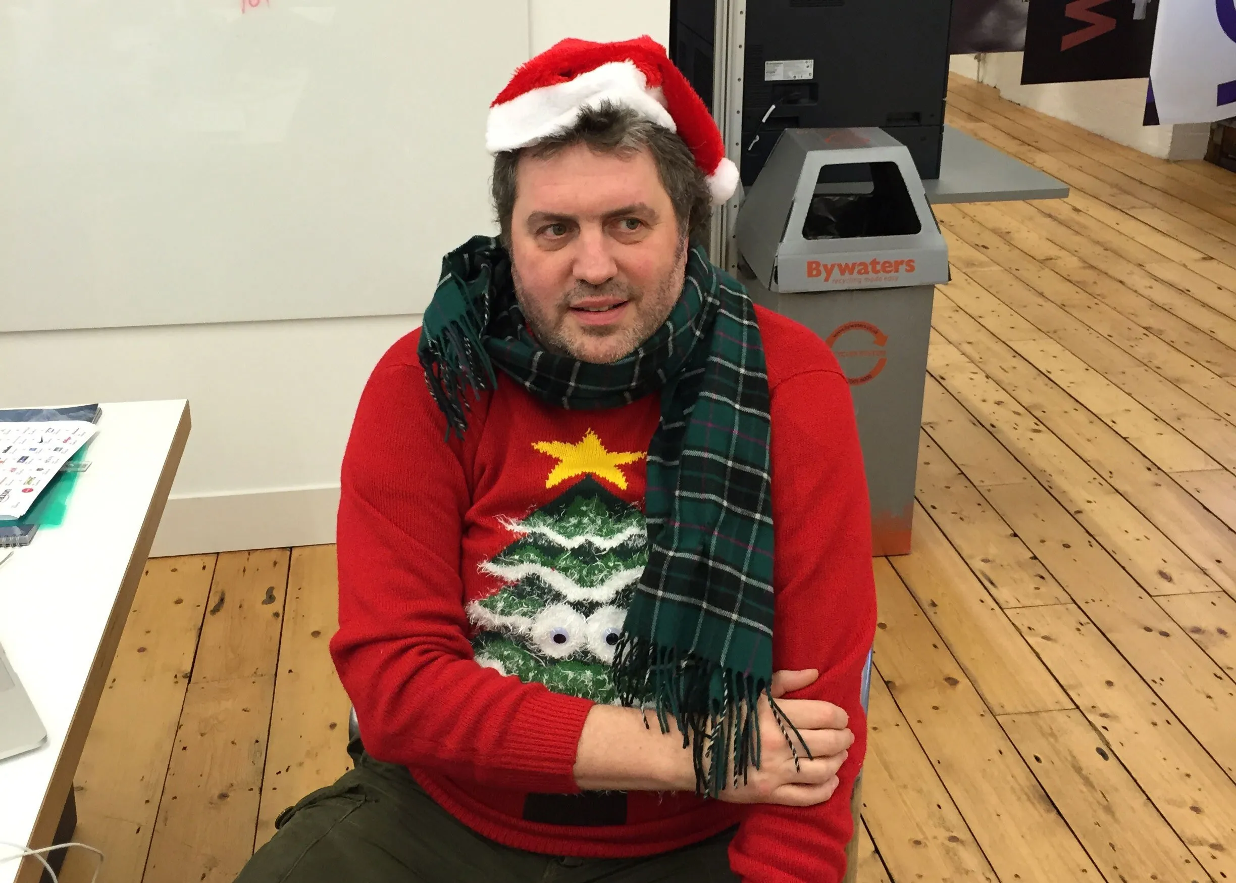 A colleague in a Santa hat, tartan scarf and red Christmas-tree jumper sits with his arms folded in the studio.
