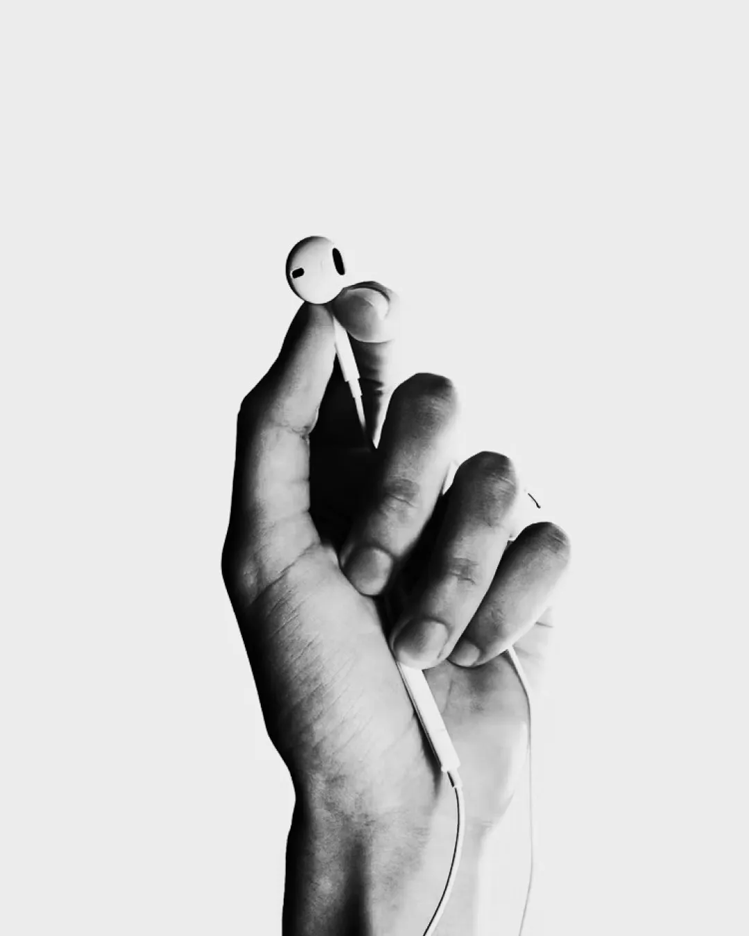 A black-and-white photo of a hand holding a white earbud by its stem against a plain white background.