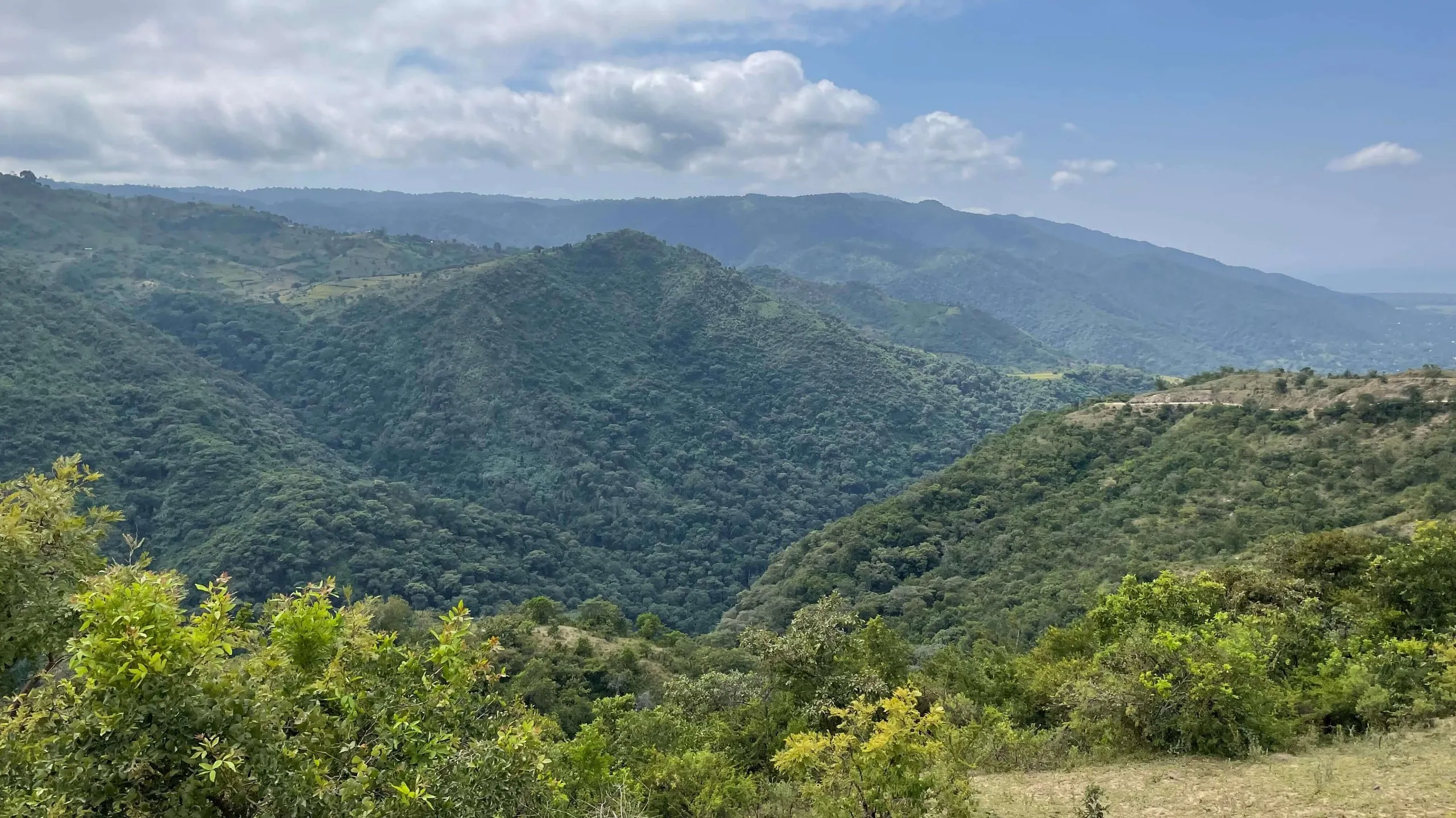 A view across steep, green hills in Tanzania under a blue sky with scattered cloud.