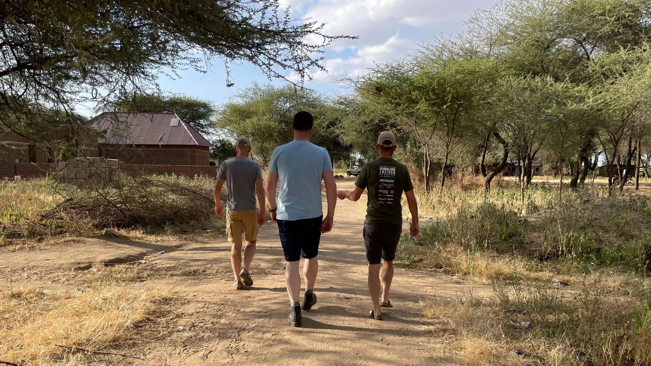 Three men walk away from the camera along a dusty track lined with acacia trees, a low building beyond them.