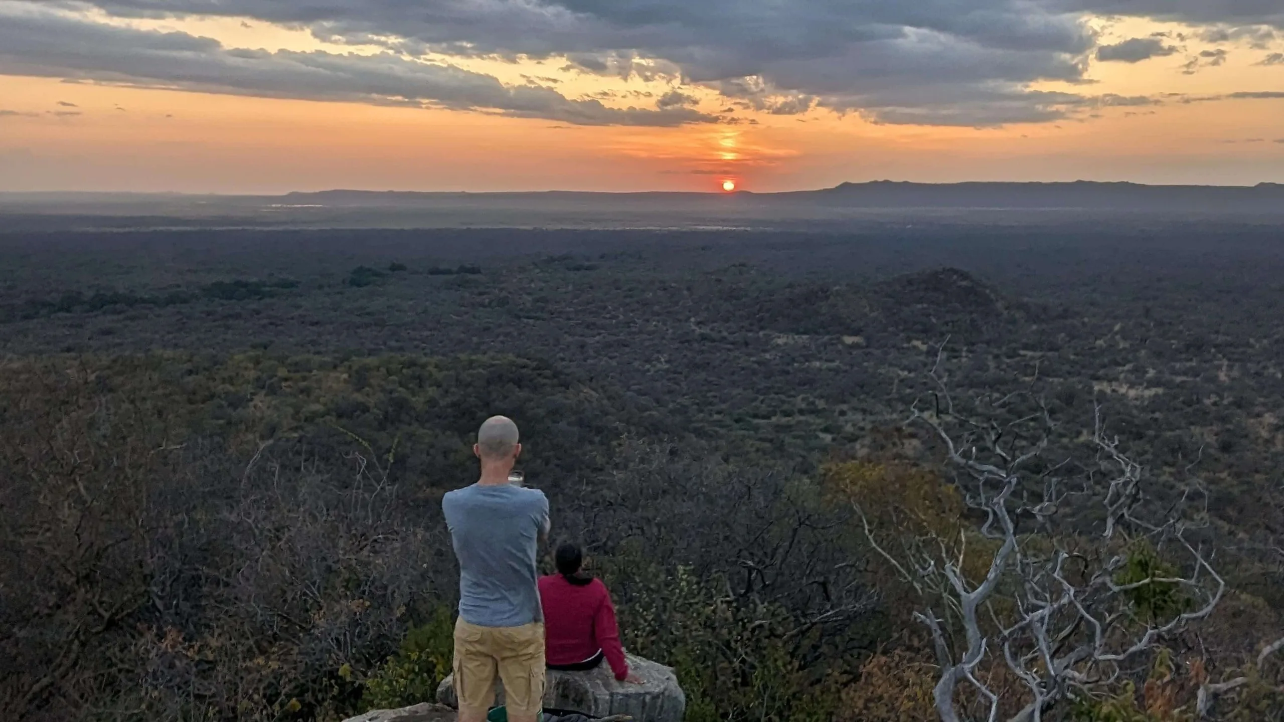 Two people sit on a rock looking out over a dark plain as the sun sets under a band of orange cloud.