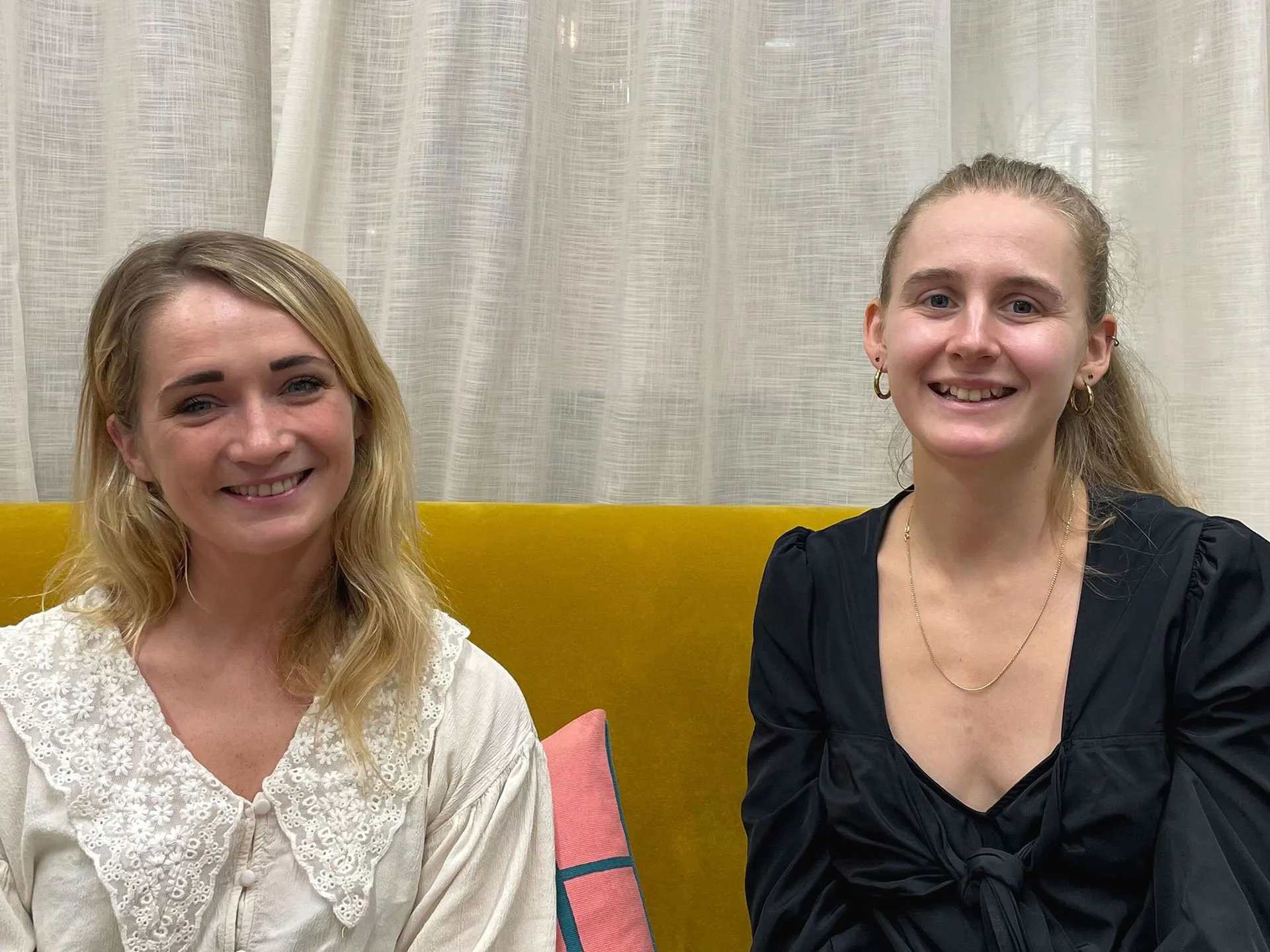 Two smiling young blonde women sitting side by side on a yellow couch in front of sheer curtains. The woman on the left wears a white lace-trimmed blouse, and the woman on the right wears a black top with gold jewelry.