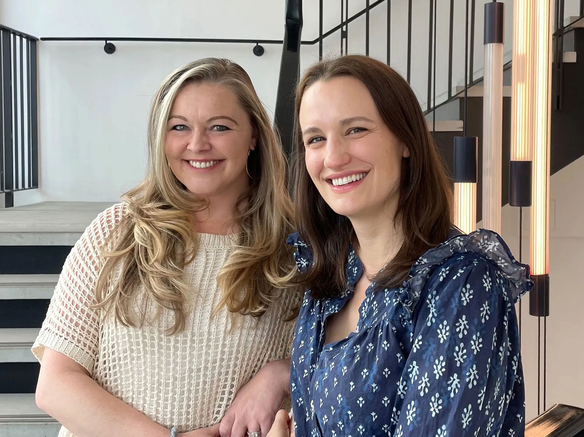 Two smiling women standing together indoors in front of a modern staircase. The woman on the left has long blonde hair and wears a cream knit top, while the woman on the right has brown hair and wears a patterned blue blouse.