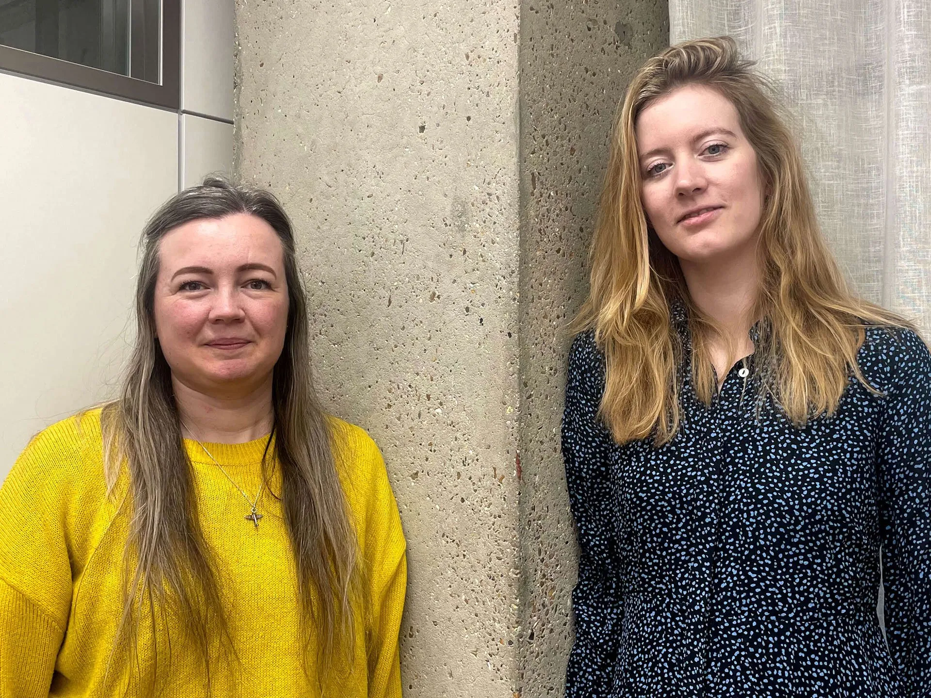 Two women standing next to a concrete pillar, with the woman on the left wearing a bright yellow sweater and the woman on the right wearing a dark patterned long-sleeve shirt.