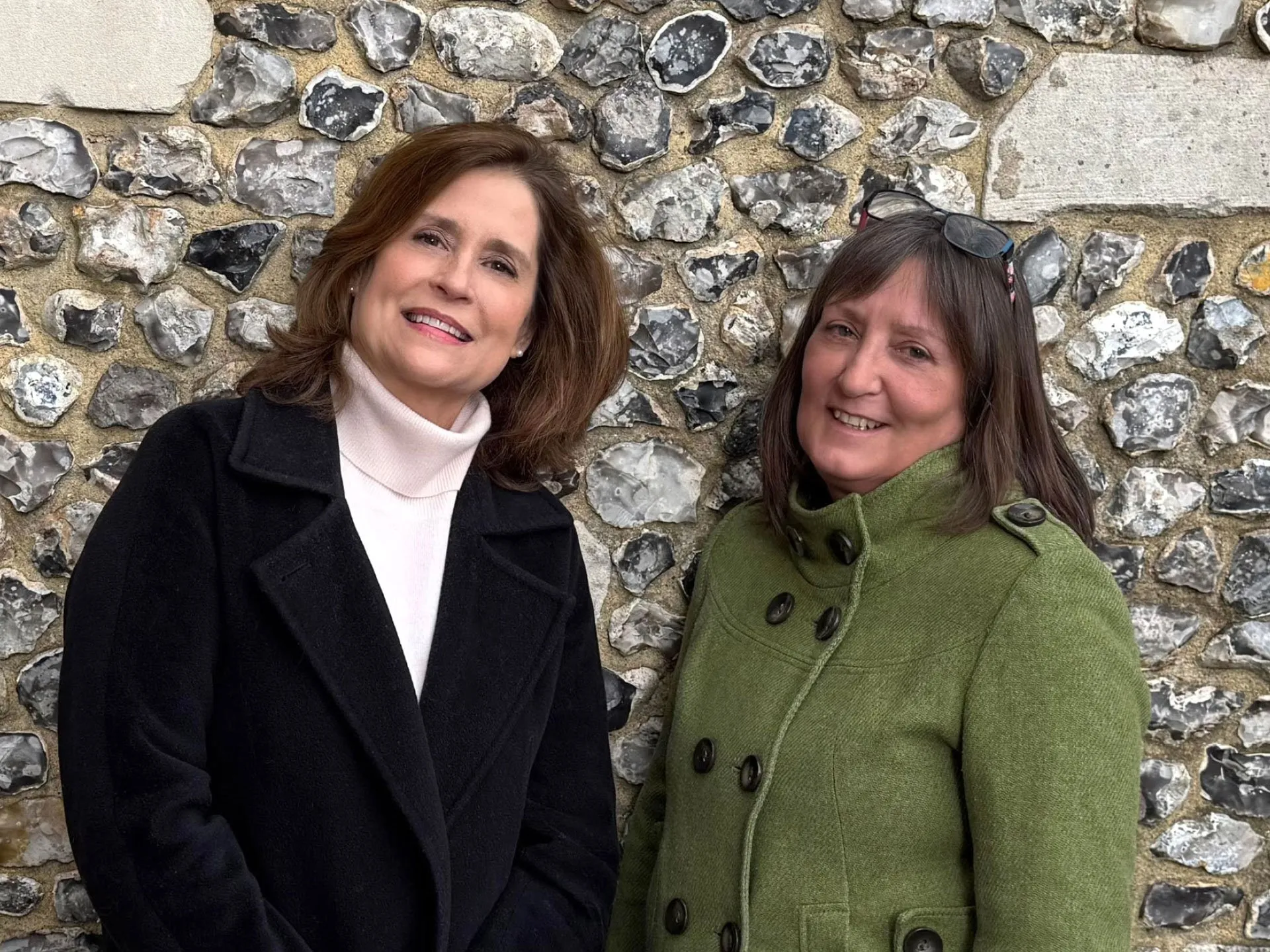 Two smiling women pose side by side in front of a flint stone wall, one wearing a black coat and white turtleneck and the other wearing a green coat with glasses on her head.
