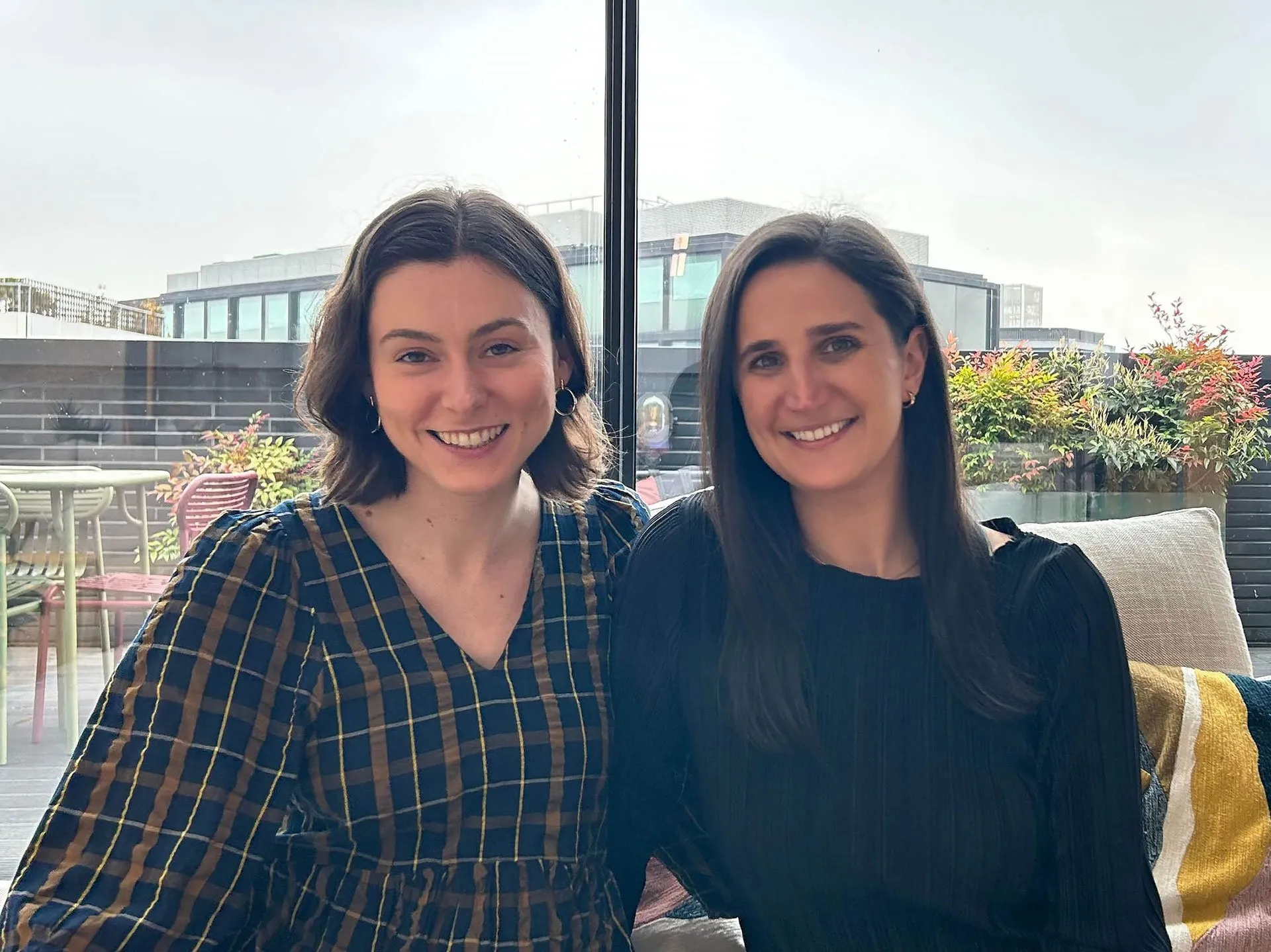 Two smiling women sit side by side indoors in front of a large window overlooking a rooftop patio with plants. The woman on the left wears a brown and blue plaid blouse, and the woman on the right wears a black top.