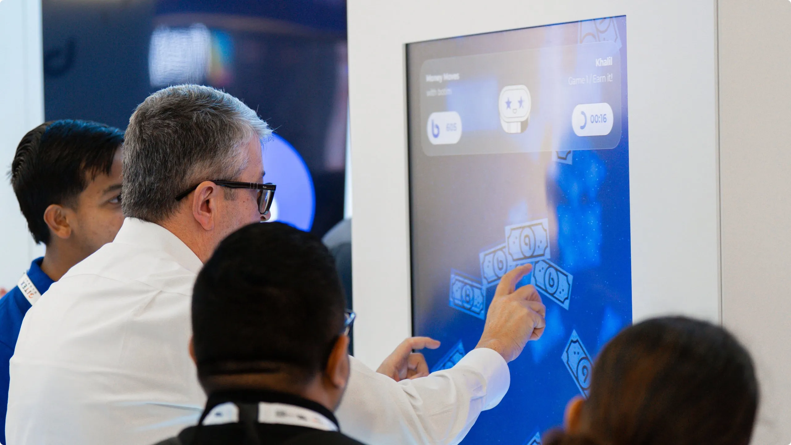 A man in a white shirt points at a large touchscreen on the botim stand while others look on; banknote graphics float across the screen.