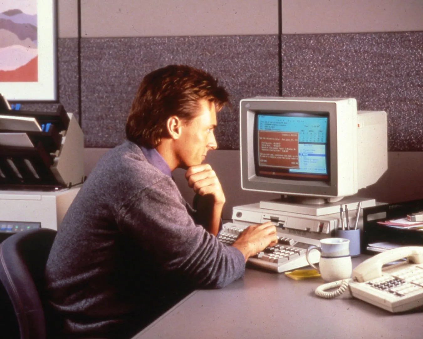 Man sitting at a desk in an office, resting his chin on his hand while looking at a vintage computer monitor and keyboard. A landline phone, mug, and printer are visible on the desk.