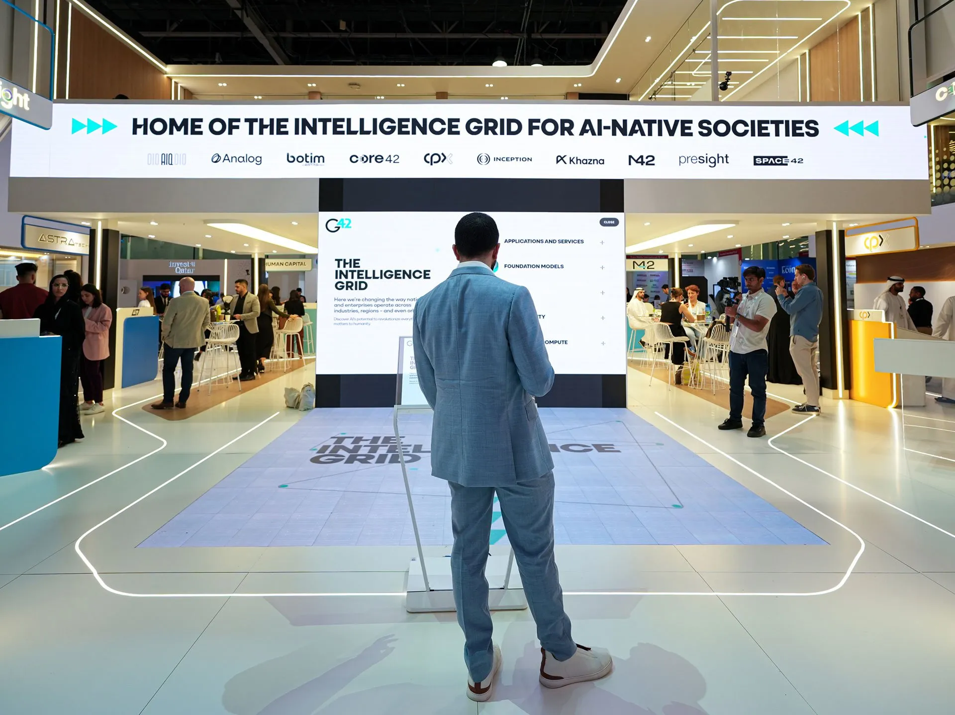 A man in a light blue suit stands in the center of a modern tech exhibition booth, facing a large vertical screen displaying an interactive presentation for G42's Intelligence Grid. Overhead, a wide illuminated sign reads, HOME OF THE INTELLIGENCE GRID FOR AI-NATIVE SOCIETIES, while other attendees mingle in the background.