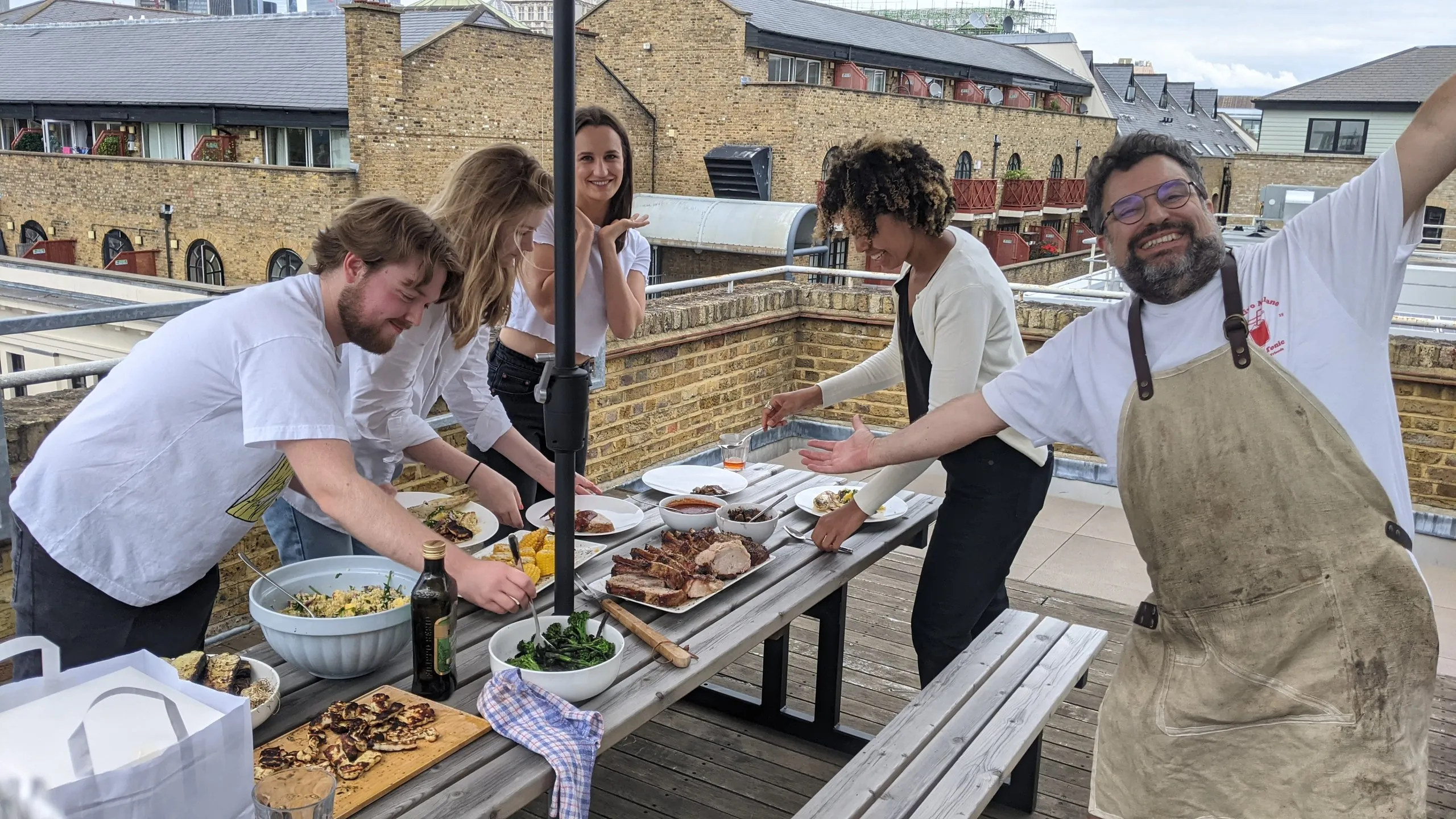 Colleagues lay out a shared lunch on a rooftop table, with bowls of salad and platters of meat, while a man in a chef's apron throws his arms out.