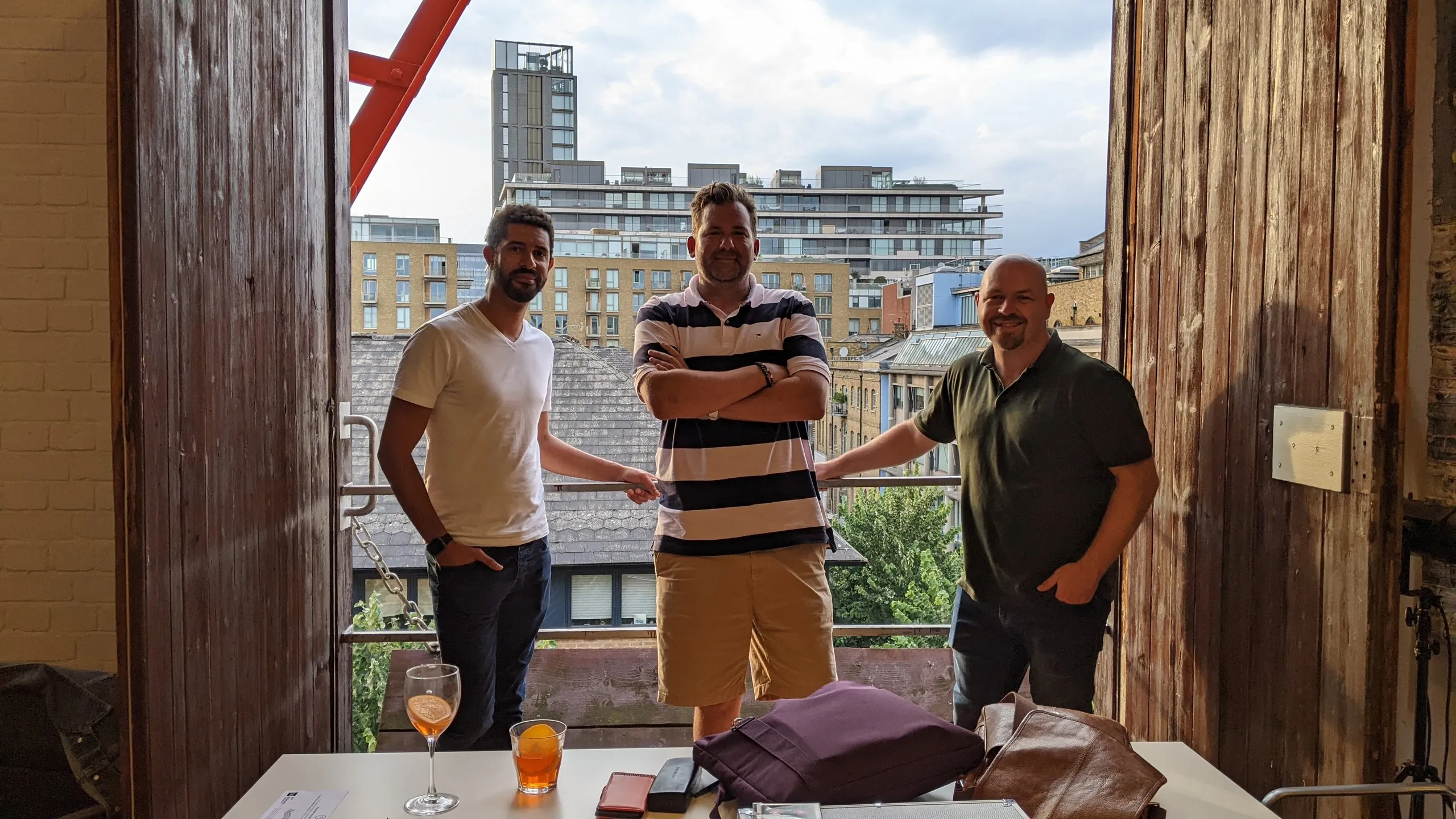 Three men stand on a balcony between the open wooden doors of the studio, with drinks on the table in front of them.