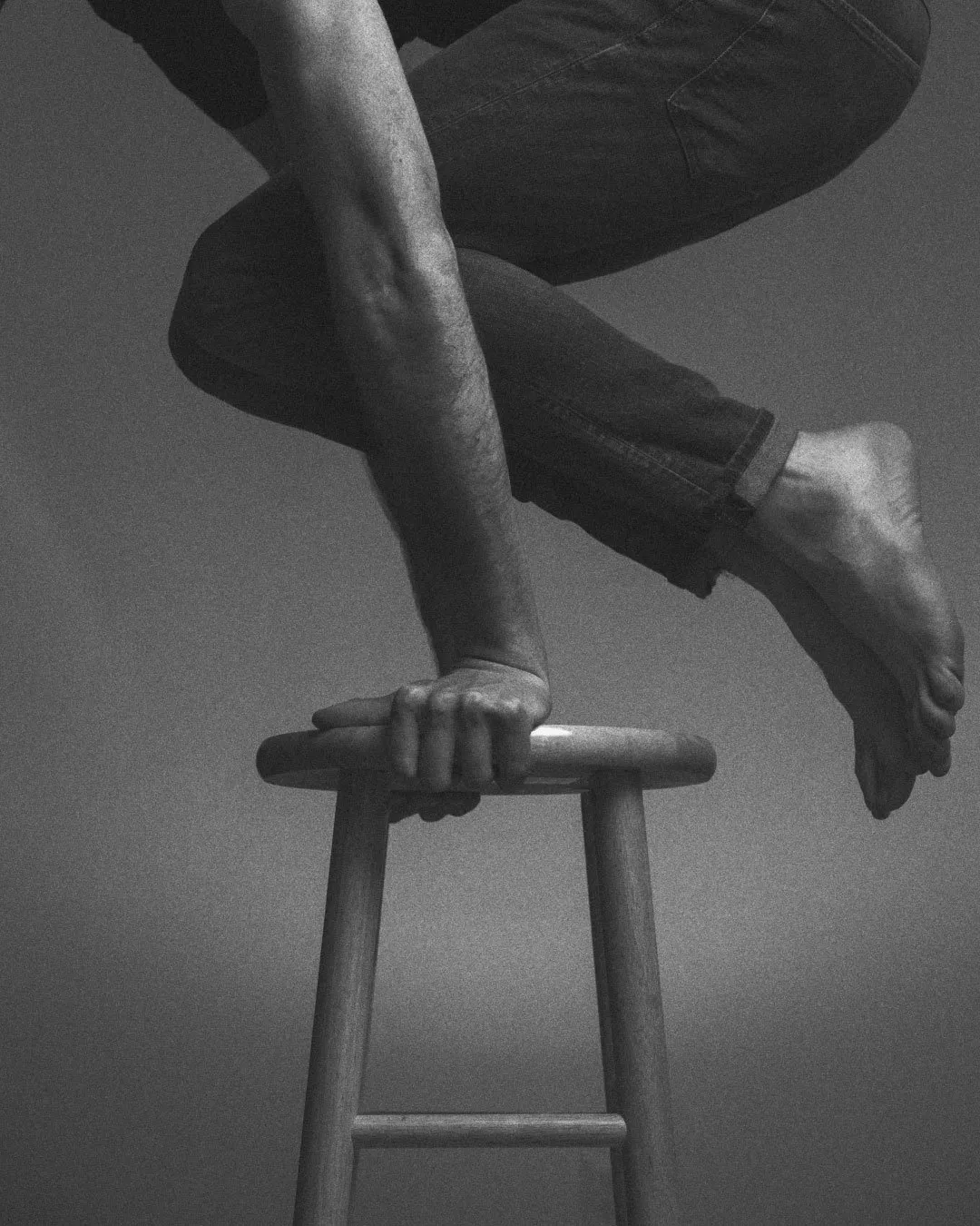 A black and white close-up of a bare arm and hand gripping the seat of a wooden stool, with legs in dark jeans bent above and a bare foot suspended in the air.
