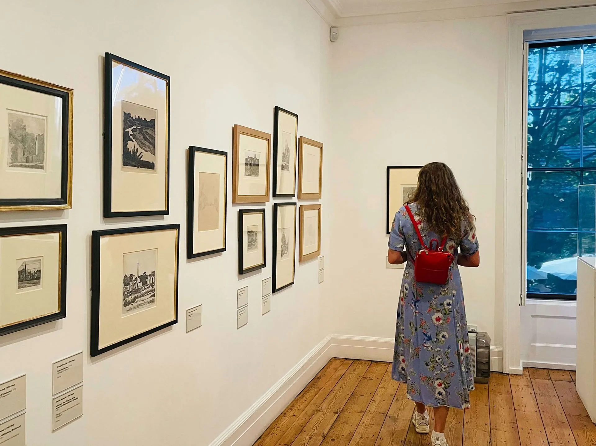 A person in a blue floral dress with a small red backpack stands in an art gallery, viewed from behind as they look at framed black-and-white prints hanging on white walls.