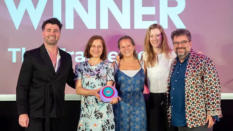 Five people on stage in front of a 'Winner' screen, one holding a pink and purple award, the man on the right in a bold geometric-patterned jacket.