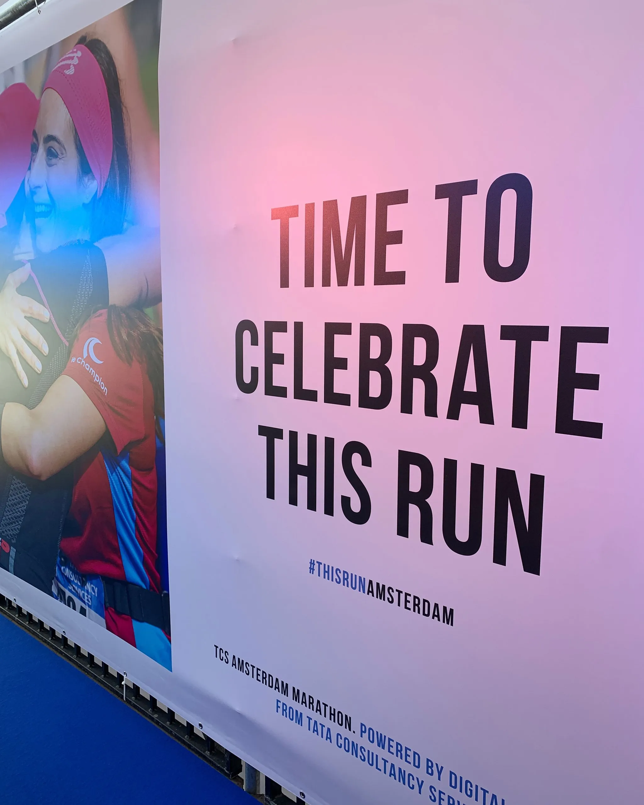 A large pink event banner reading "TIME TO CELEBRATE THIS RUN" in bold black letters above the hashtag #THISRUNAMSTERDAM, with smaller text for TCS Amsterdam Marathon Powered by Digital, and a photo on the left of two women runners hugging.