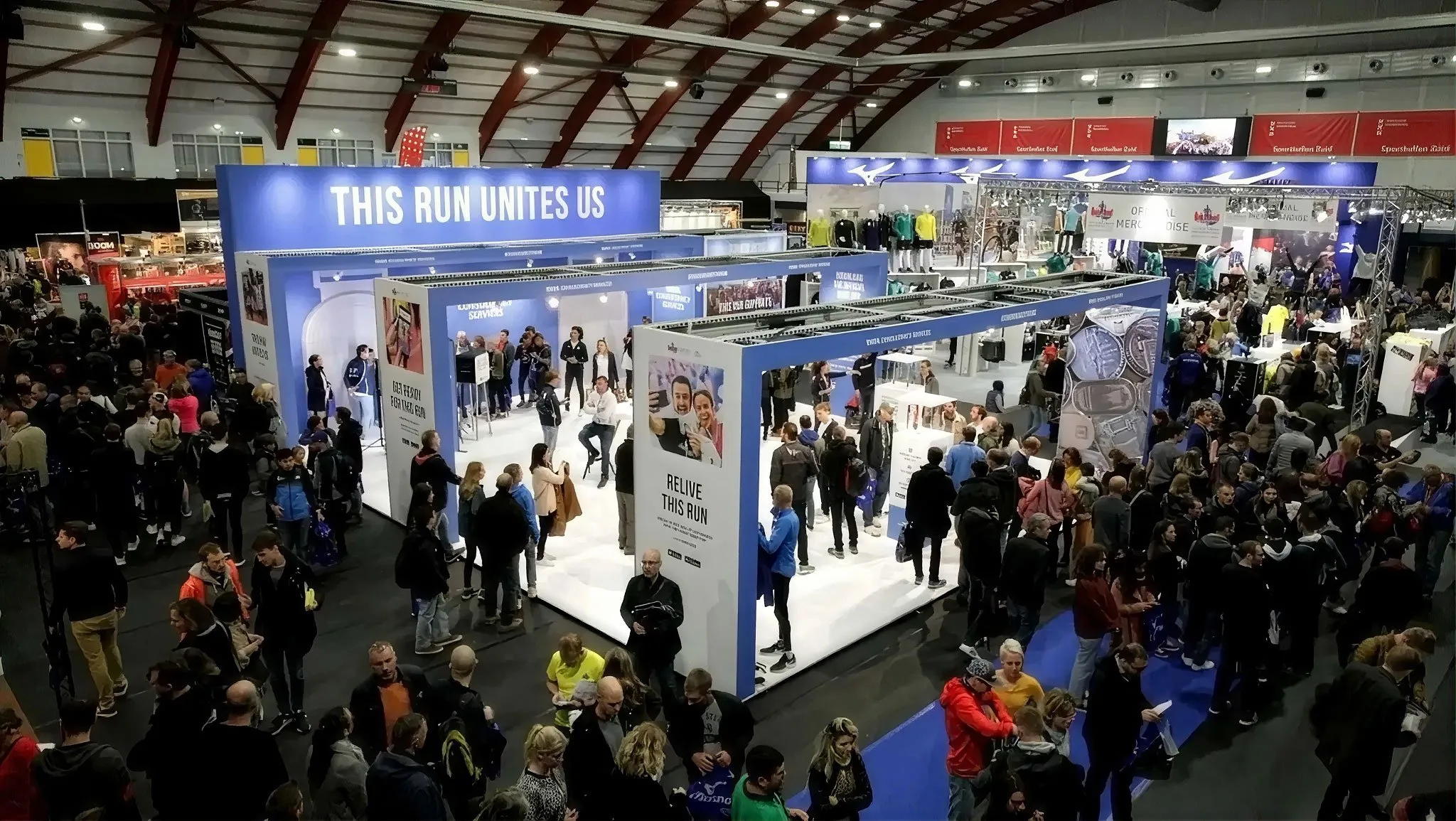 Crowded indoor exhibition hall with a large blue booth reading THIS RUN UNITES US and many attendees walking between displays.