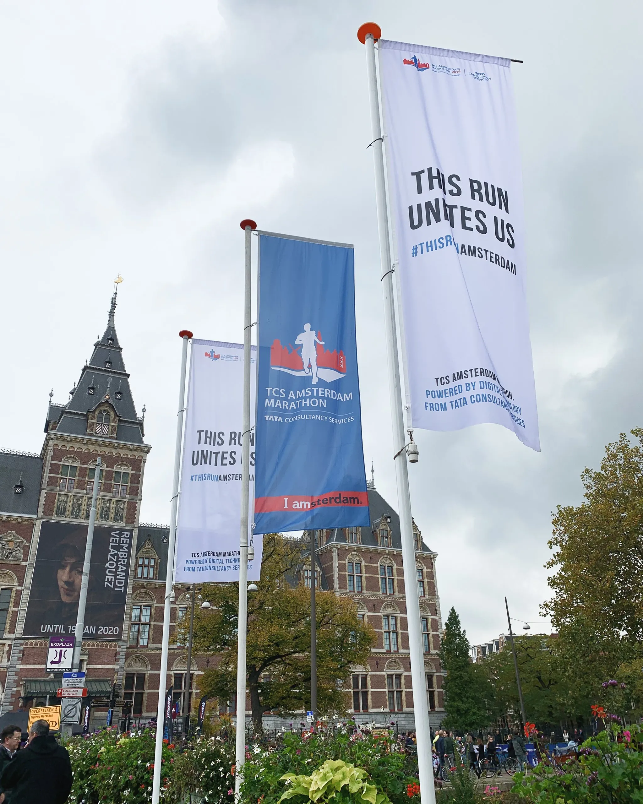Three tall banners for the TCS Amsterdam Marathon stand in front of the Rijksmuseum under a cloudy sky. The white banners read “This Run Unites Us,” and the blue banner shows a runner silhouette with “I amsterdam.” A large museum banner advertises a Rembrandt–Velázquez exhibition, with flower beds and pedestrians below.
