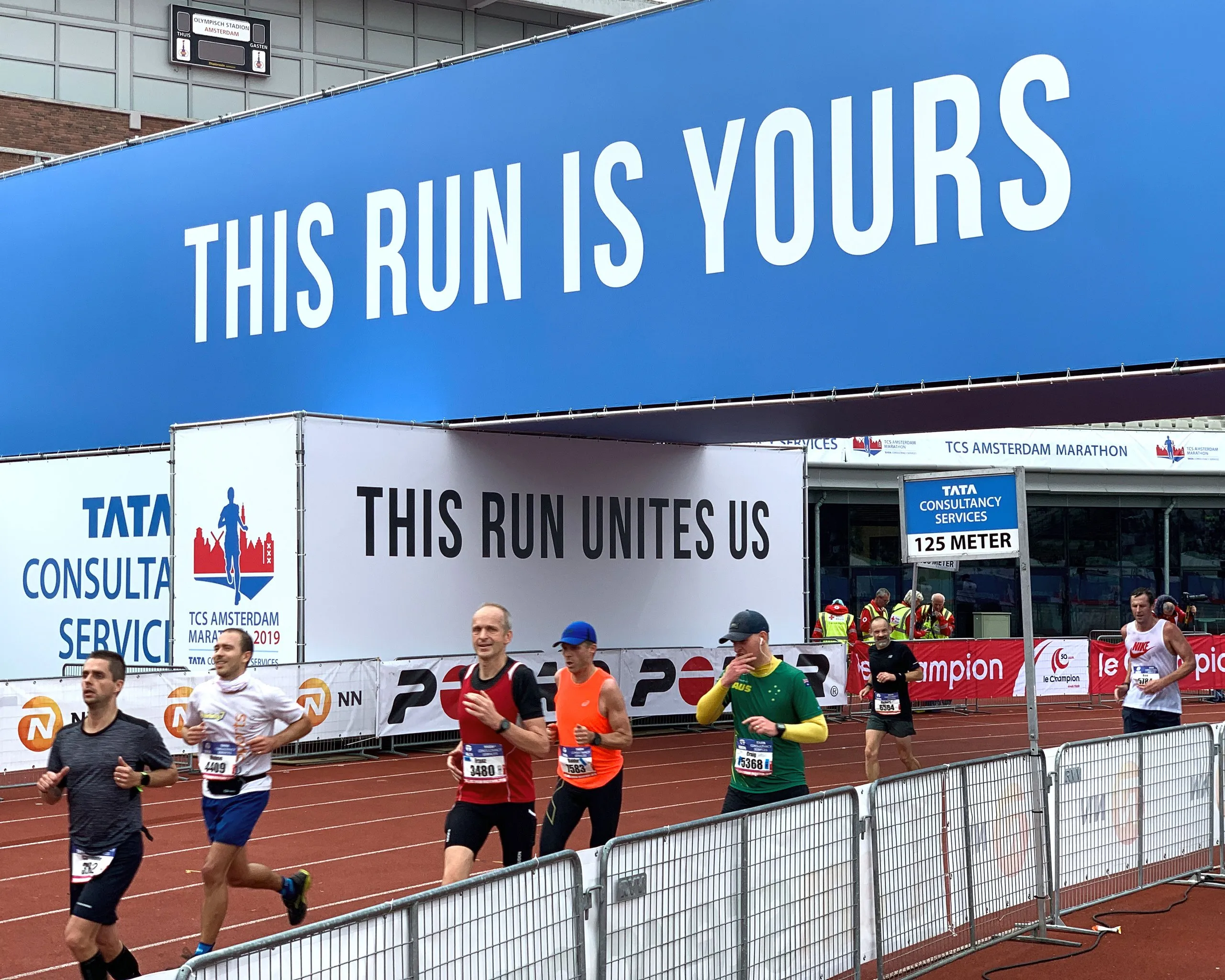 Runners pass a large blue banner reading THIS RUN IS YOURS and a white banner reading THIS RUN UNITES US at the TCS Amsterdam Marathon.
