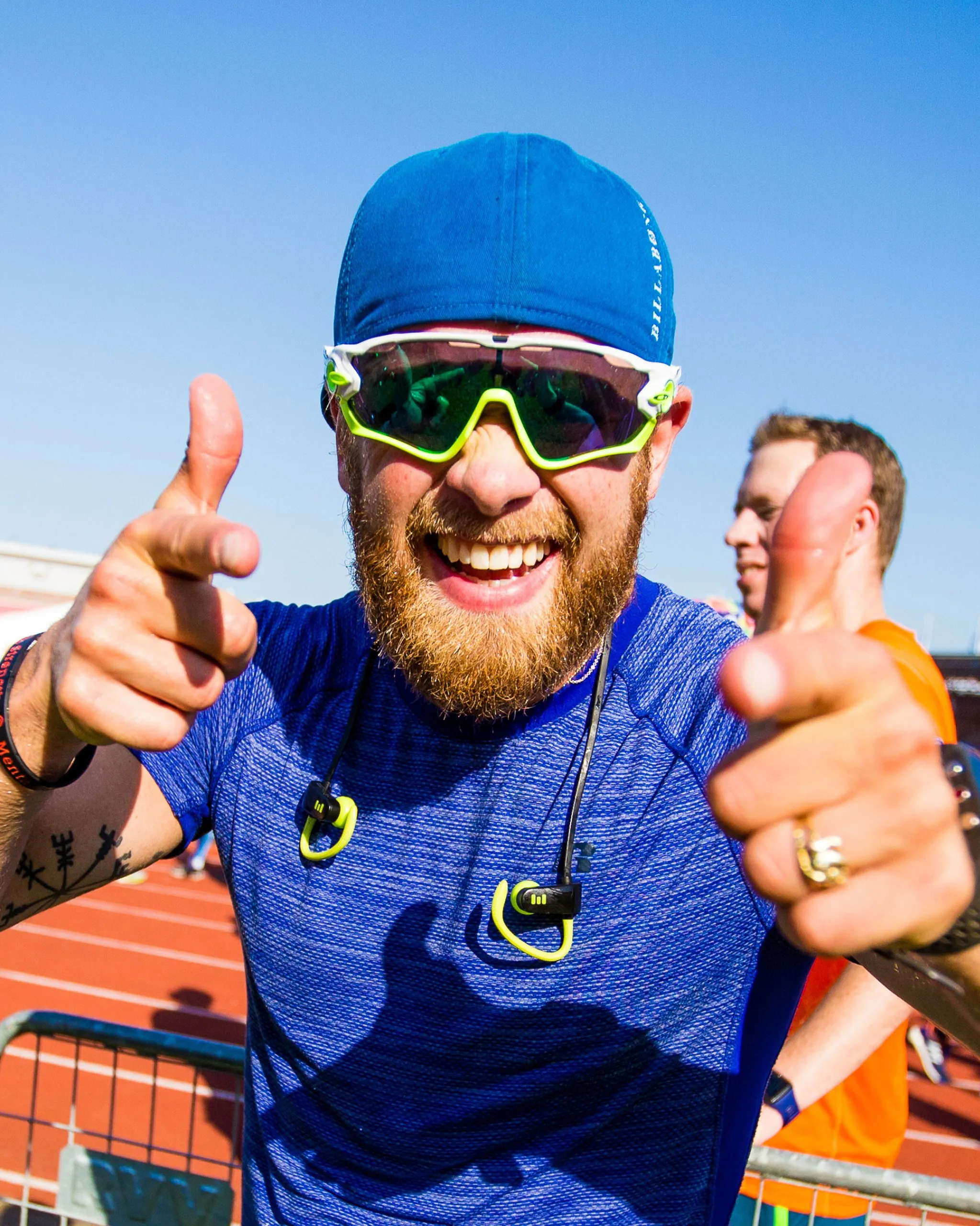 A smiling bearded man in a blue cap, blue athletic shirt, and neon sunglasses gives two thumbs up on a sunny outdoor track.