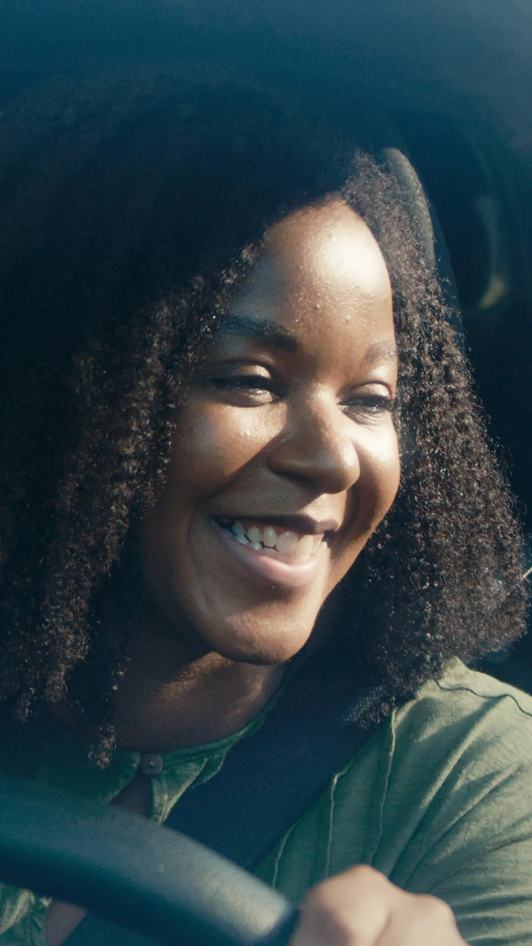 A woman at the wheel of a car, smiling in the sunlight.